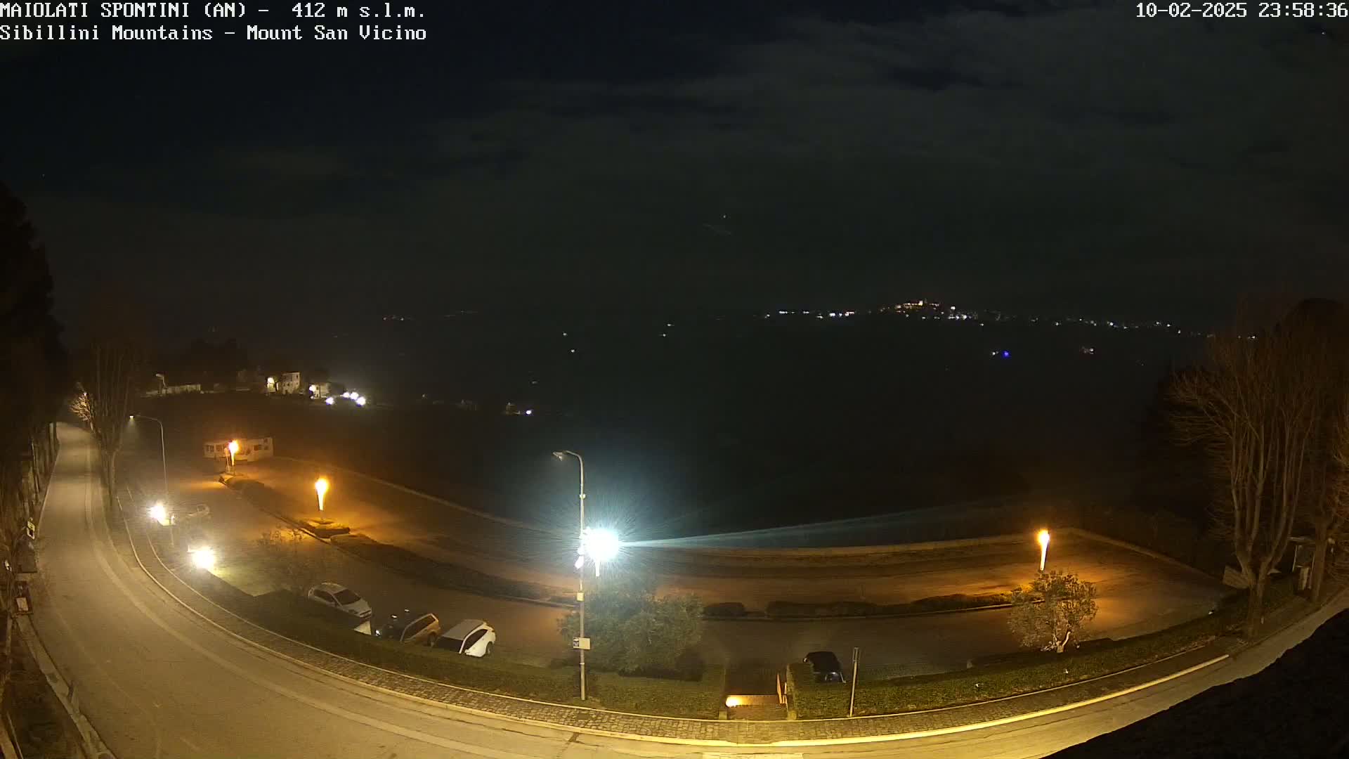 A nighttime view of a curving road illuminated by streetlights, a parking area with several cars, and a dark body of water in the distance under a mostly clear sky.