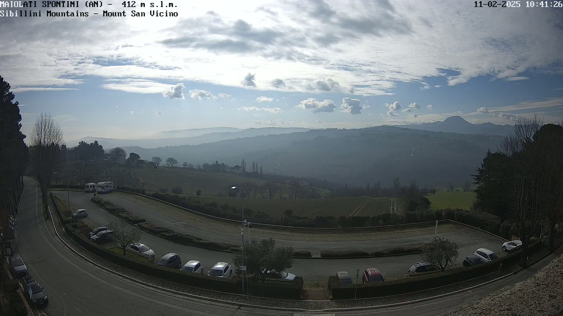 A partly cloudy sky overlooks a parking lot with several cars and two vans, curving roads, and a hazy view of distant hills and mountains.