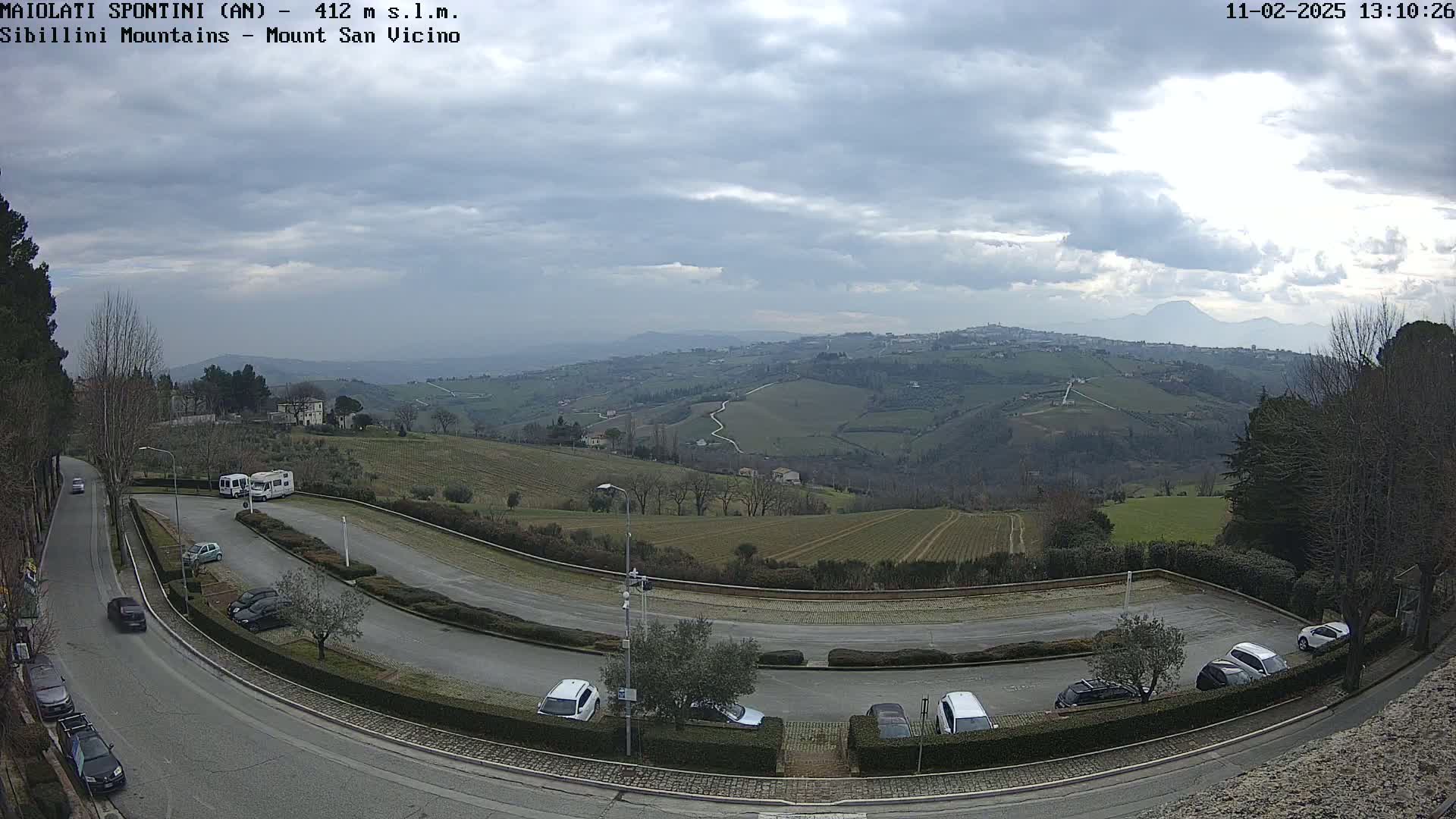 An overcast sky overlooks a parking lot and winding road nestled among rolling green hills and distant mountains.