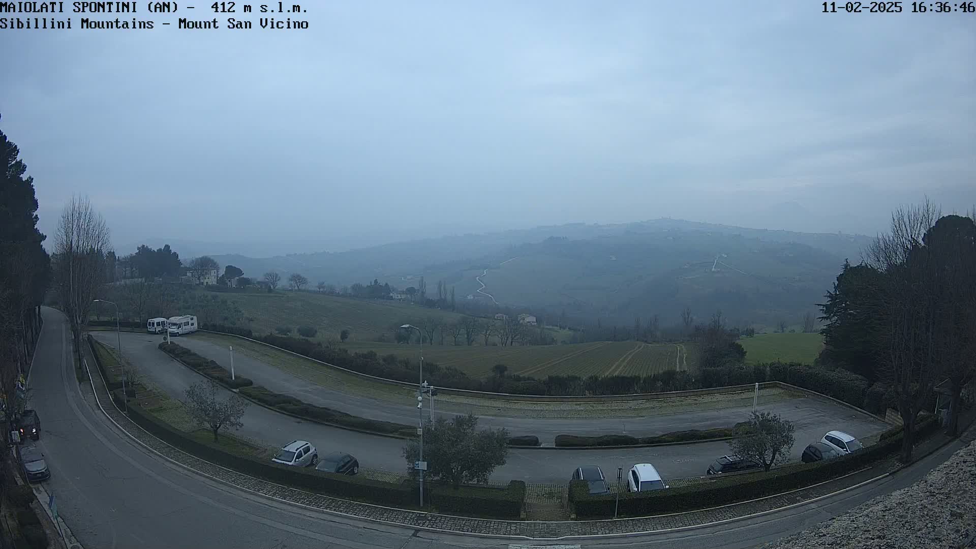 A hazy, overcast view overlooks a parking lot with several cars and RVs, situated next to a winding road that leads toward rolling hills and vineyards under a pale sky.