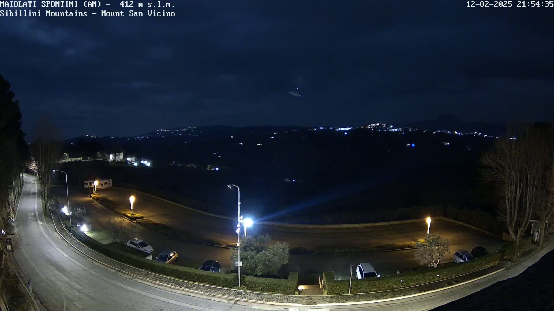 A nighttime, overhead view of a curving road with parked cars, illuminated by streetlights, overlooks a dark, hilly landscape with distant, faintly lit buildings under a mostly clear sky.