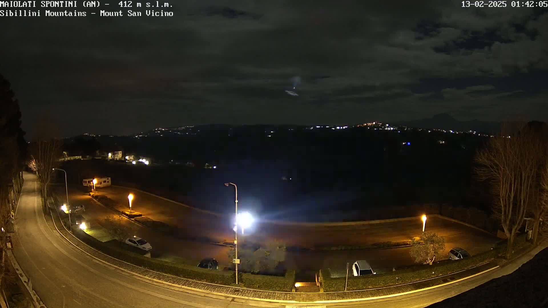 A nighttime, overcast scene shows a curving road illuminated by streetlights, with several cars parked in a lot, and distant lights from a town or city in the background.