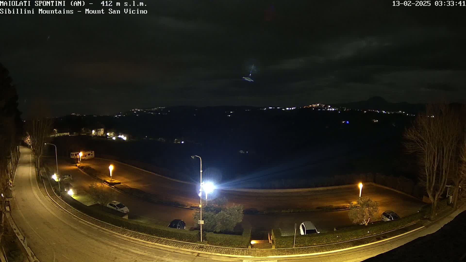A nighttime view of a curving road and parking area, illuminated by streetlights, with a distant, dimly lit town and hills under a cloudy sky.