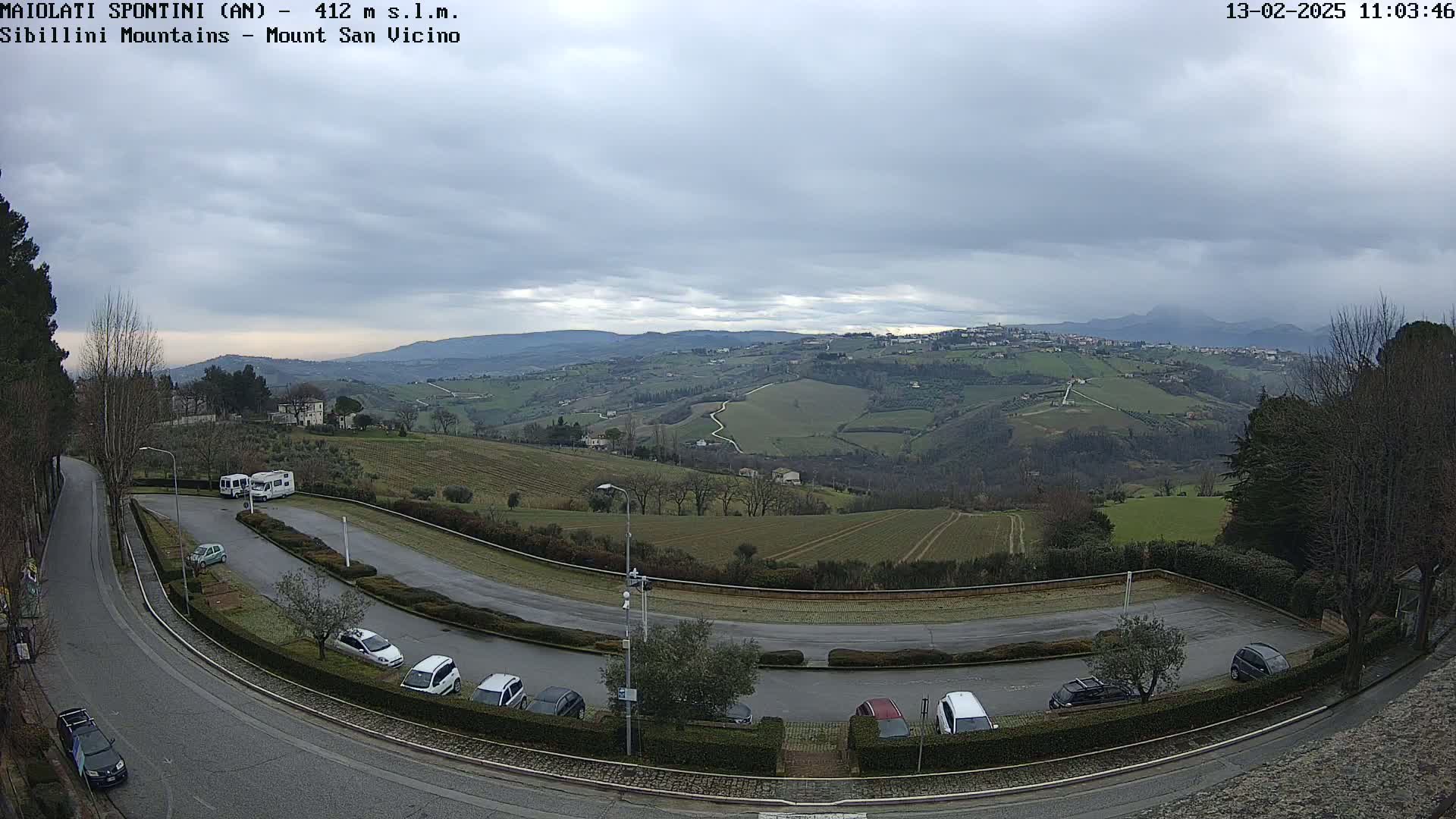 A mostly cloudy day overlooks a parking lot situated on a curved road, with a hilly, green landscape and distant mountains in the background.