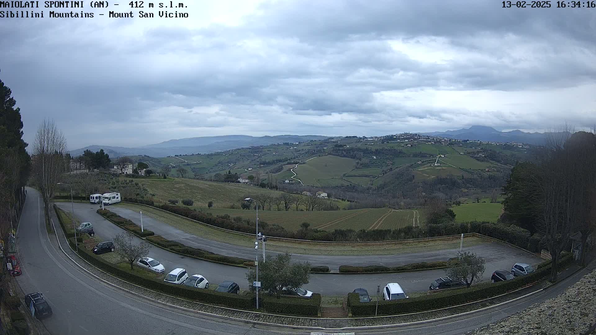 A mostly cloudy day reveals a view of rolling hills with vineyards and a parking lot containing several cars and RVs, overlooked by a winding road.