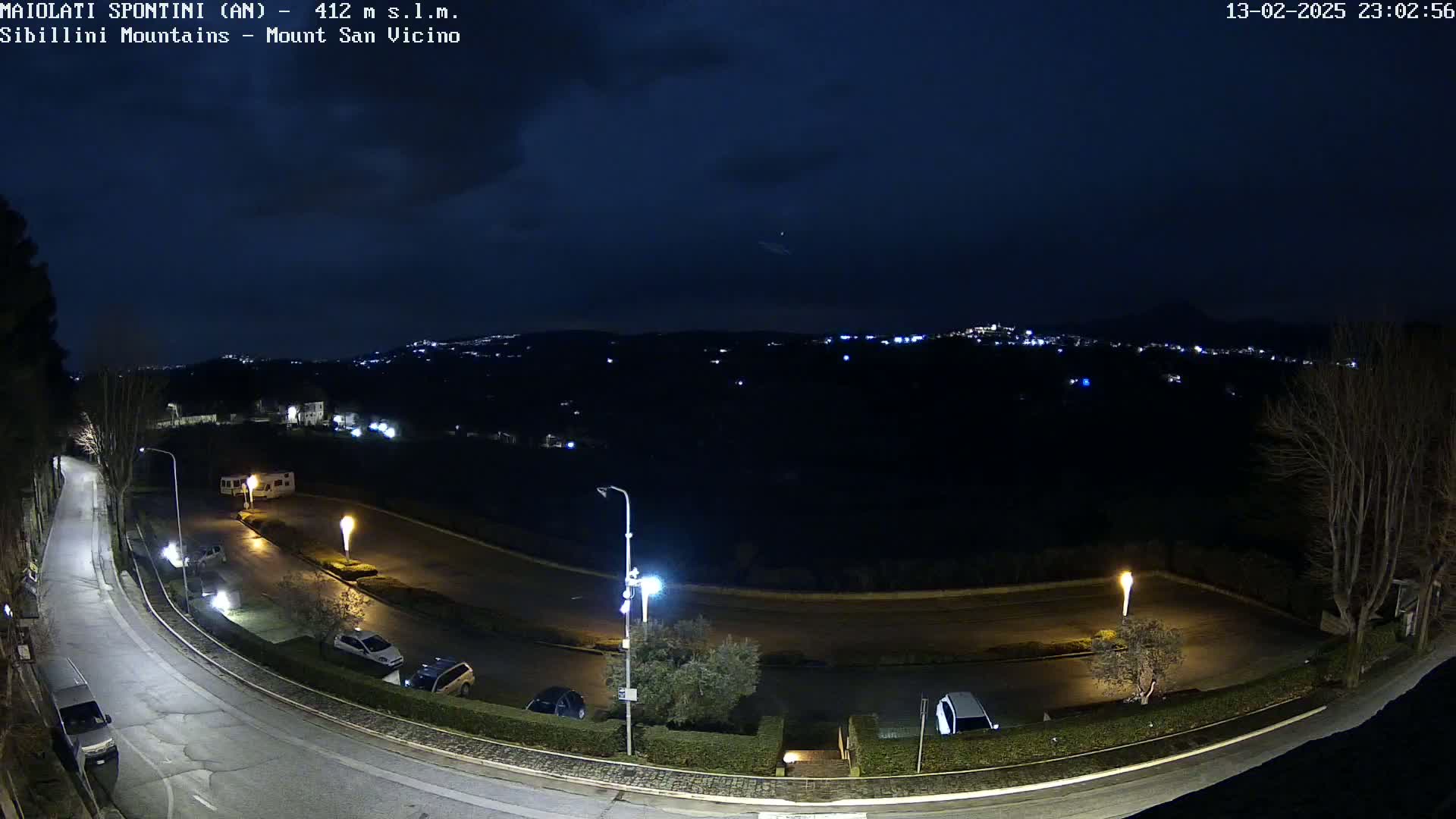 A nighttime, mostly clear view of a curving road with parked cars and a distant city glowing under a dark sky.
