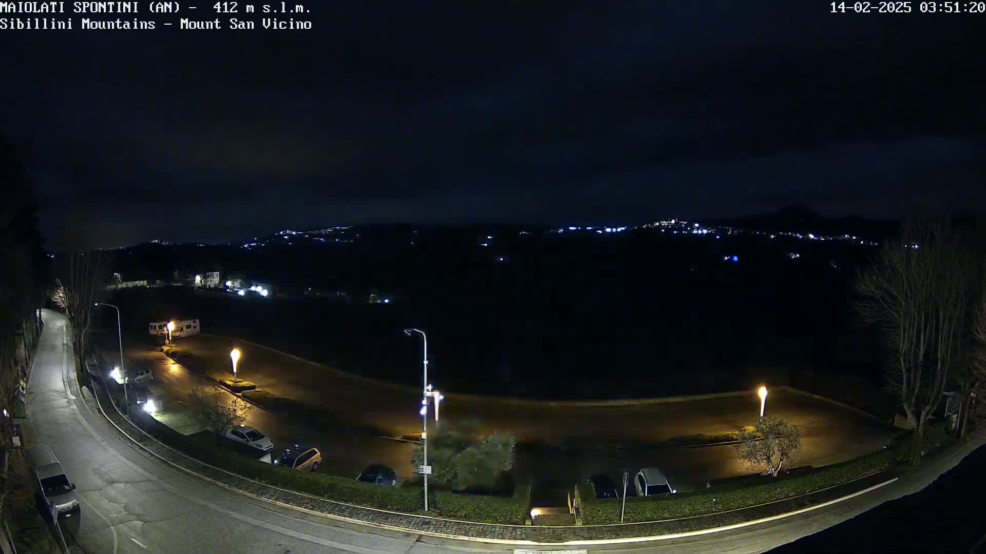 A nighttime, high-angle view of a curving road with parked cars and illuminated lampposts, overlooking a dark, sparsely lit landscape under a clear night sky.