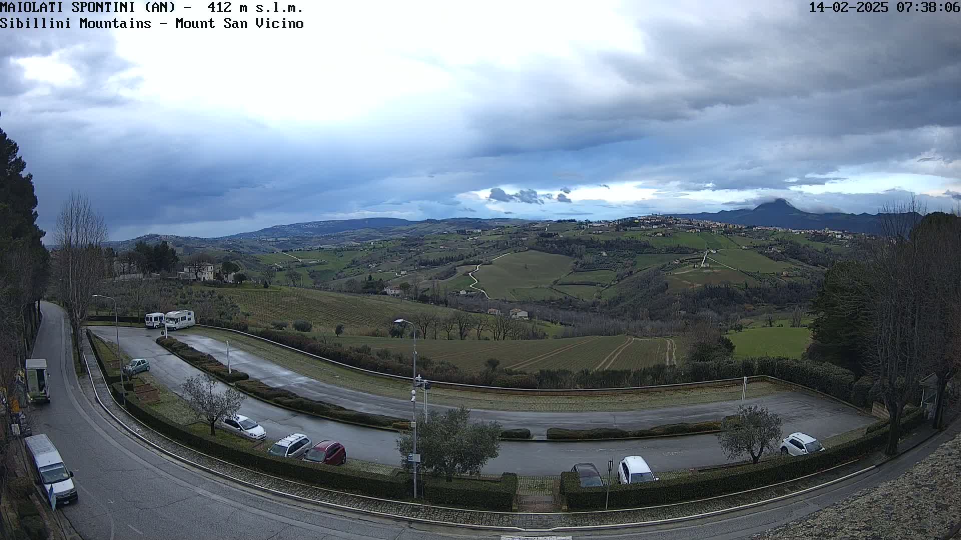 A winding road curves through a parking lot overlooking a hilly landscape under a cloudy sky, with several cars and RVs parked.