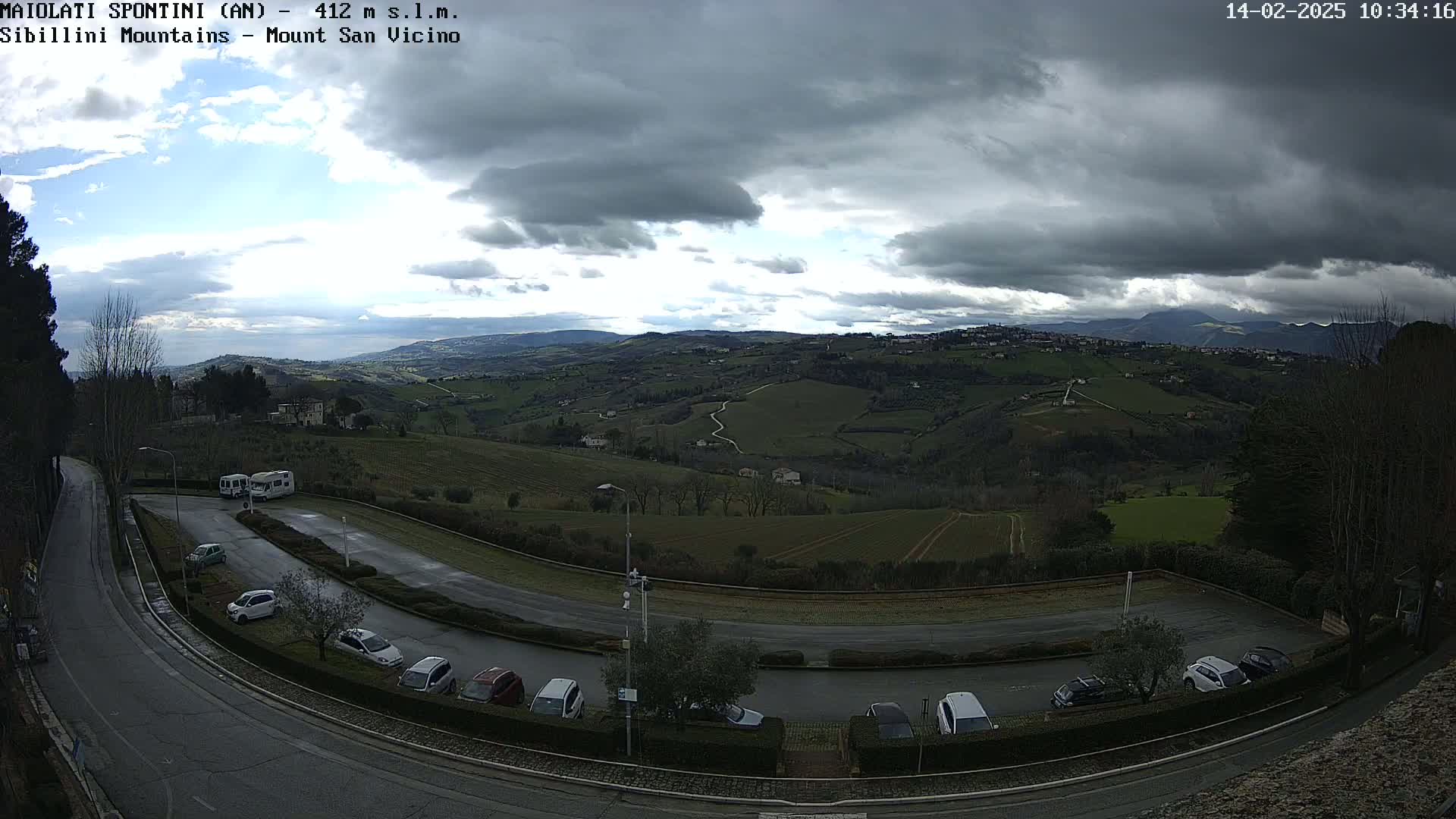 A mostly cloudy day overlooks a hilly landscape with a parking lot and road curving through the foreground, a body of water visible in the far distance.