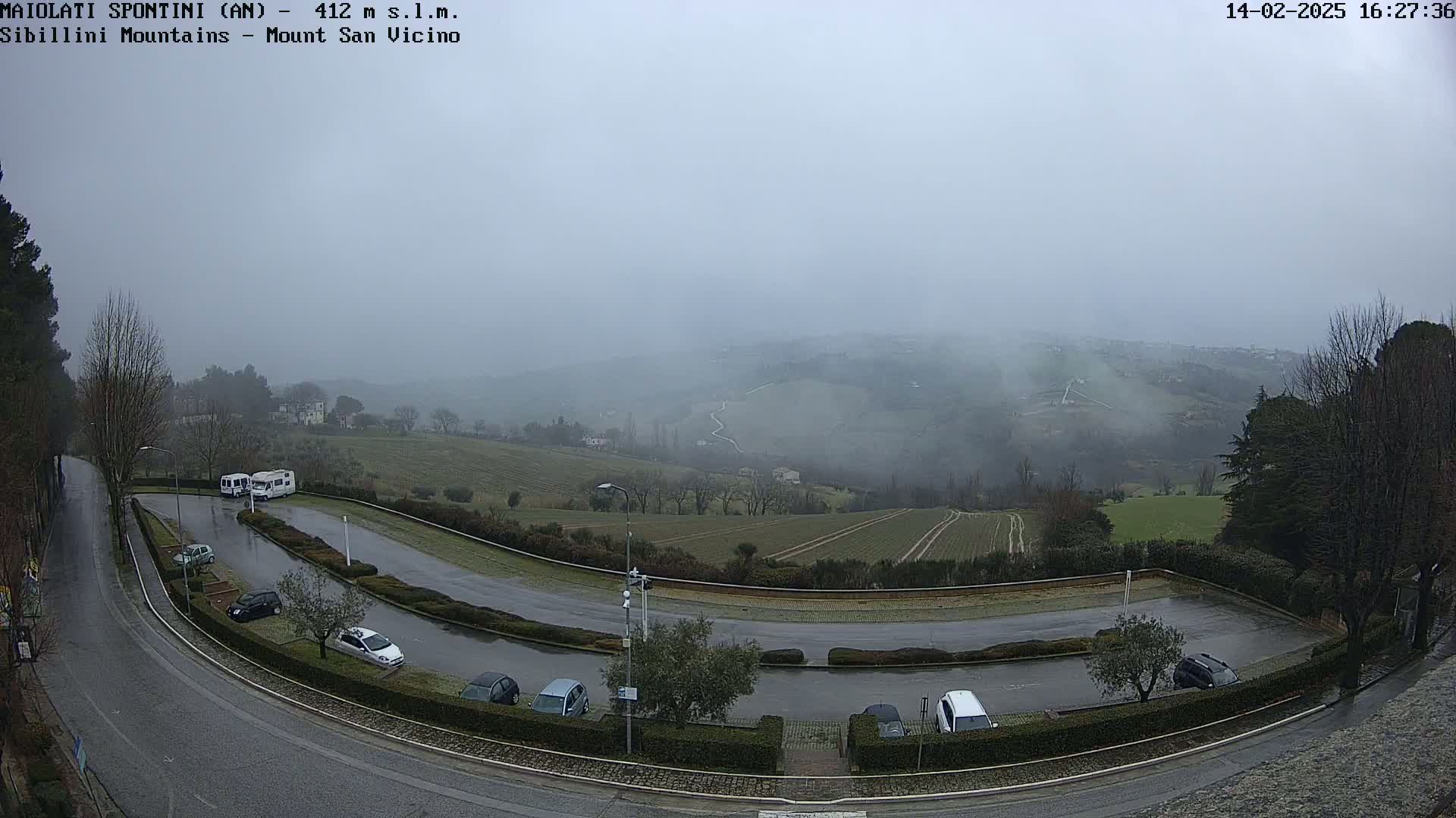 A wet, foggy view overlooking a parking lot with several cars and two campervans, and distant hills in the background.