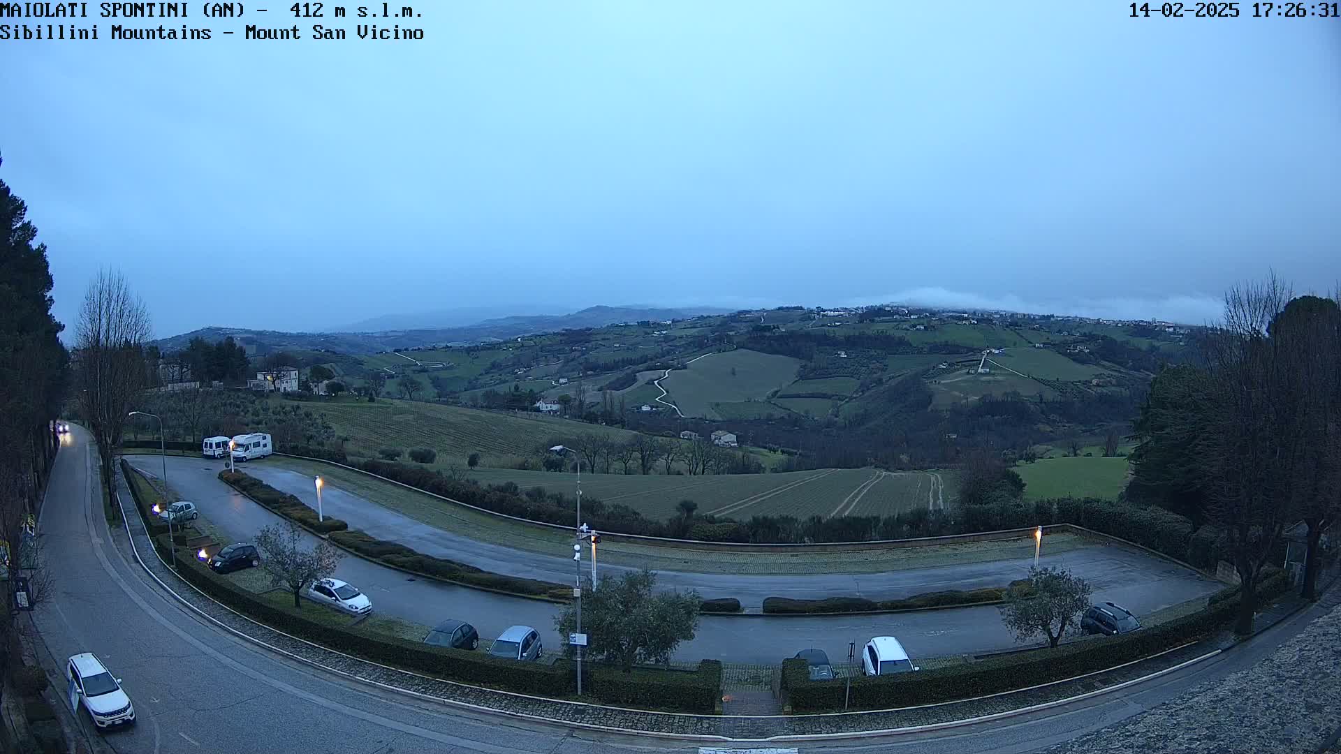 A wet road curves through a parking lot overlooking a valley of rolling hills and low-lying fog under a dusky sky.