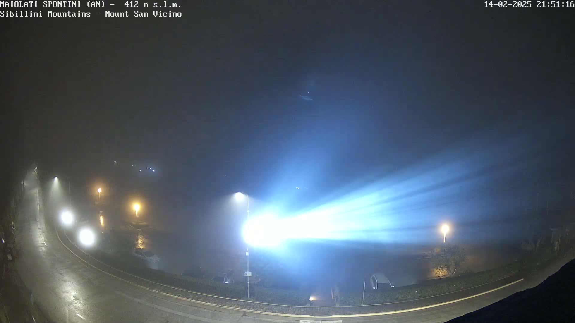 A bright light shines across a foggy, nighttime scene of a curved road and a body of water, with a few vehicles visible and streetlights illuminating the mist.