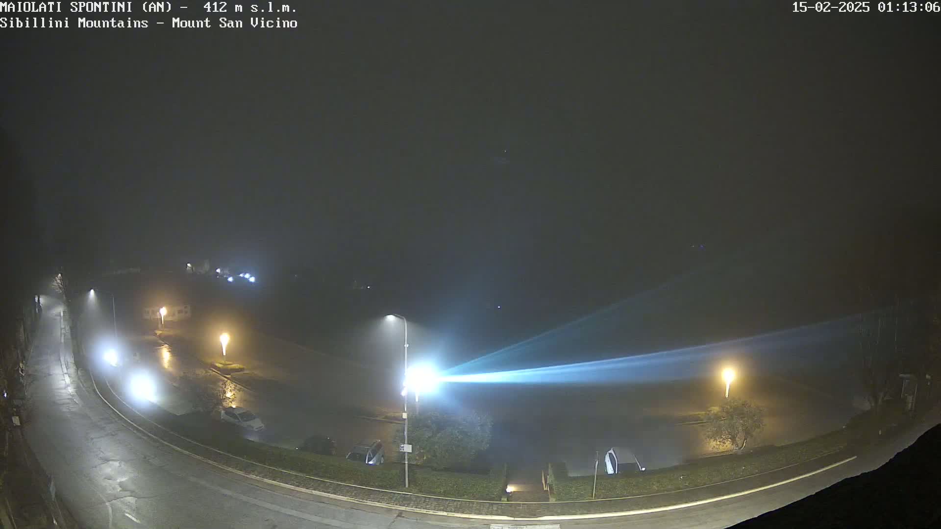 A foggy night scene shows a curved road with parked cars and streetlights illuminating a flooded parking lot, with a bright light beam stretching across the area.