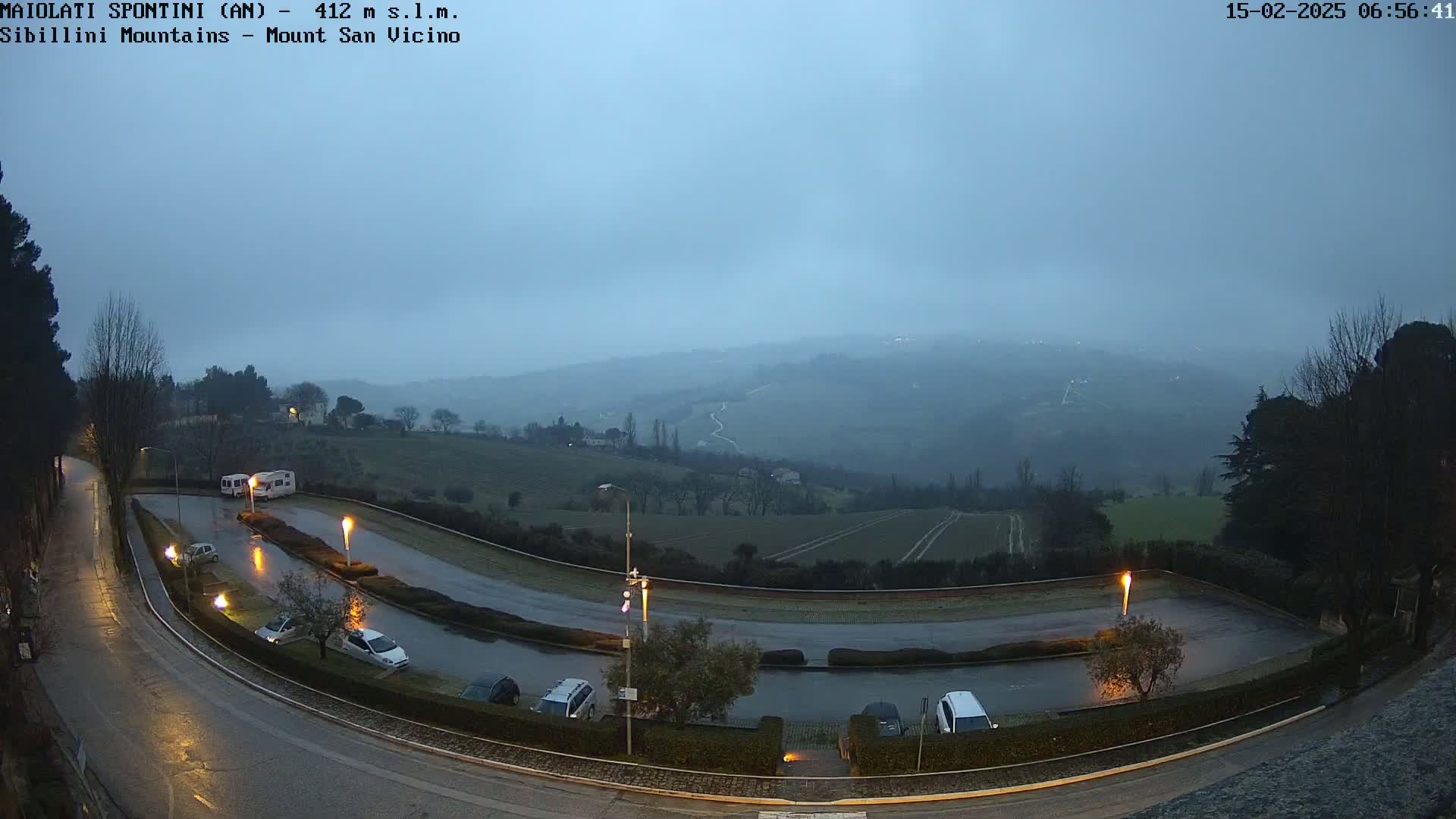 A wet, empty parking lot curves around a small hill overlooking a misty, hilly landscape at dawn.