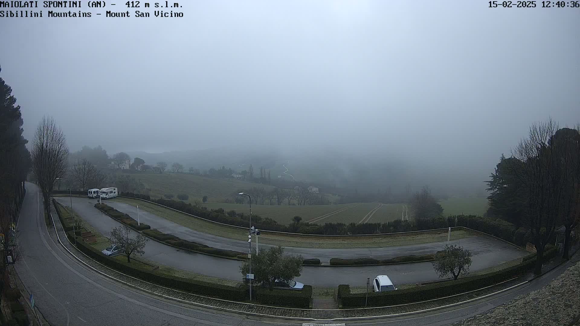 A foggy view of a winding road and parking lot with a few vehicles, overlooking a valley of farmland under a gray sky.