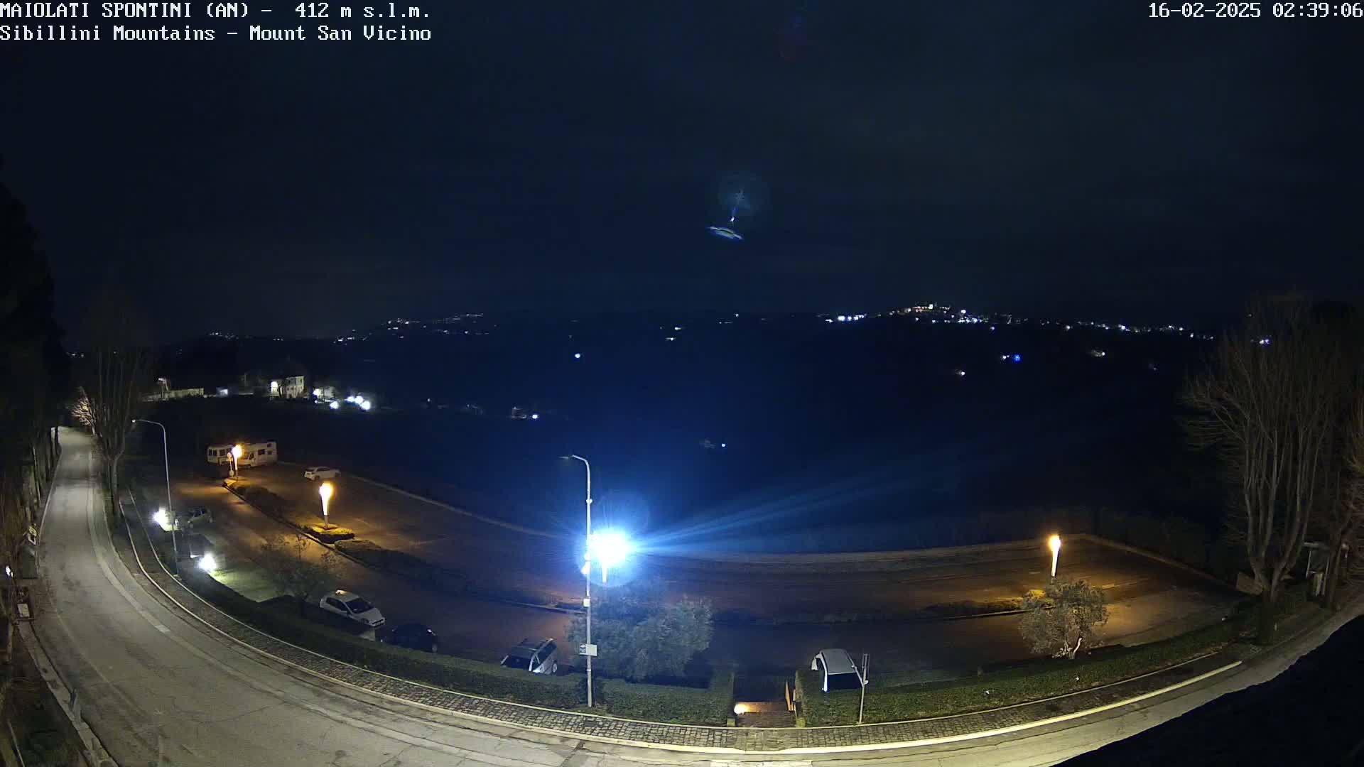 A nighttime, high-angle view of a curving road with parked cars and illuminated streetlights, overlooking a dark, distant town under a clear night sky.