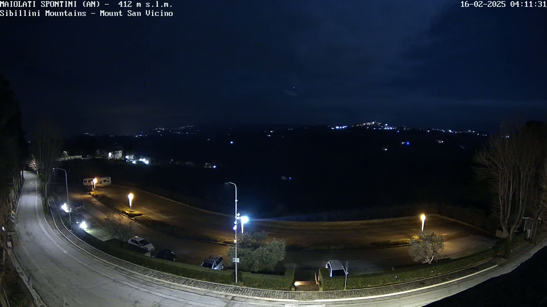 A nighttime, high-angle view shows a curving road with parked cars and streetlights, overlooking a dark, hilly landscape speckled with distant lights under a partly cloudy sky.