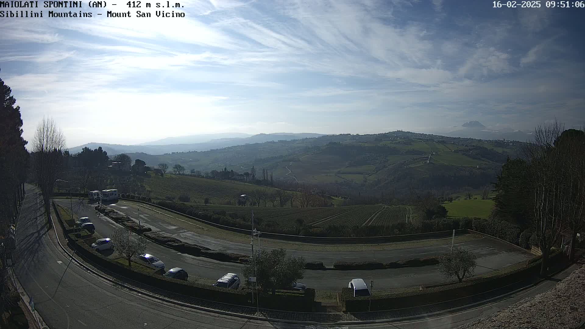 A mostly sunny day reveals a hilly landscape with vineyards and a parking lot filled with cars, overlooked by a mountain in the distance.