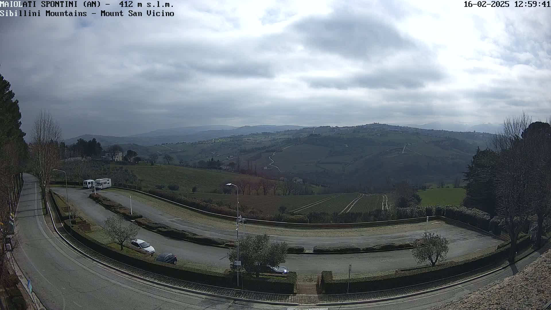A winding road curves through a parking lot overlooking a hilly landscape under a cloudy sky.
