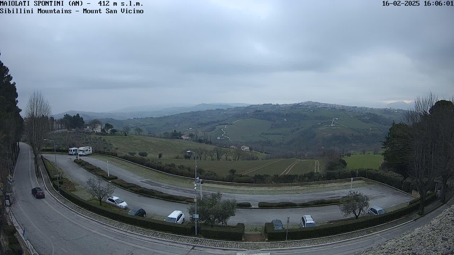 A winding road curves through a parking lot overlooking a valley of rolling green hills under an overcast sky.