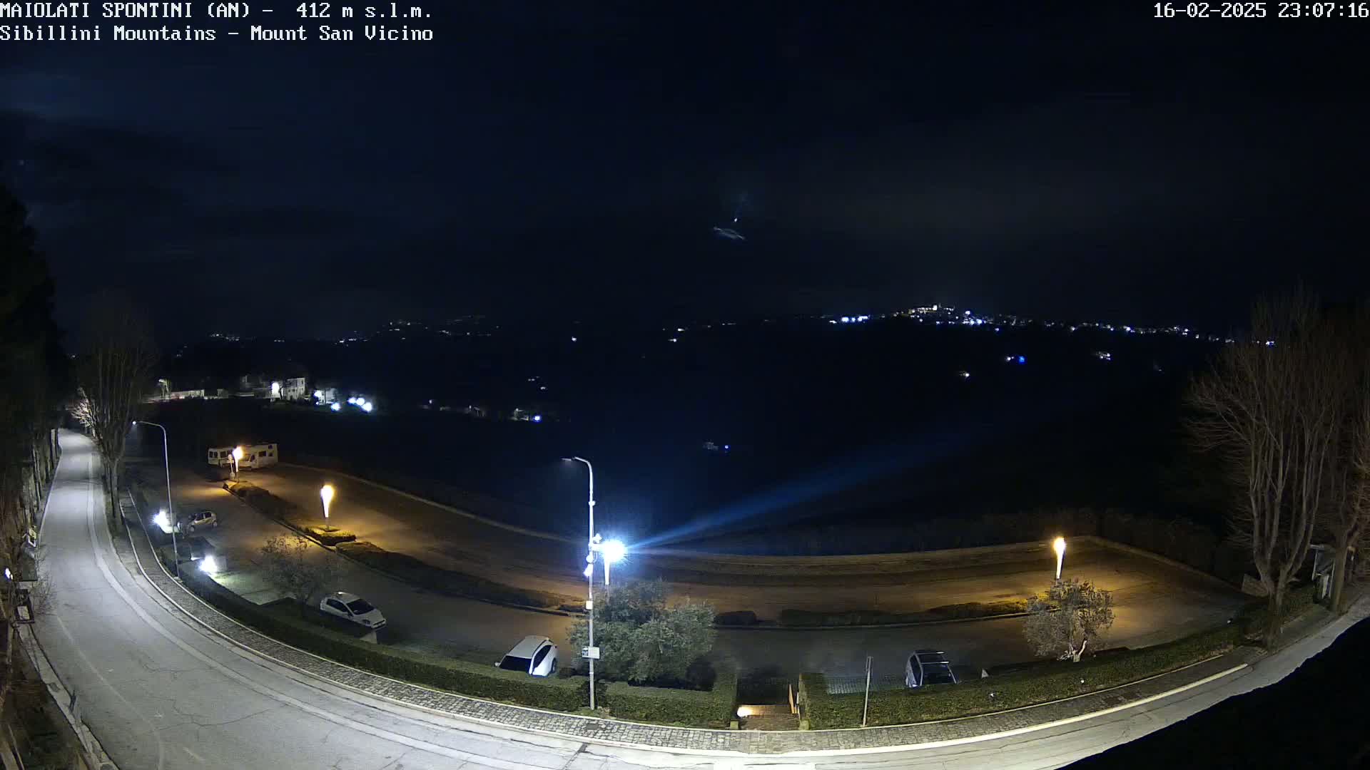 A nighttime, high-angle view of a curving road with parked cars and illuminated lampposts, overlooking a distant town with faint lights under a clear, dark sky.