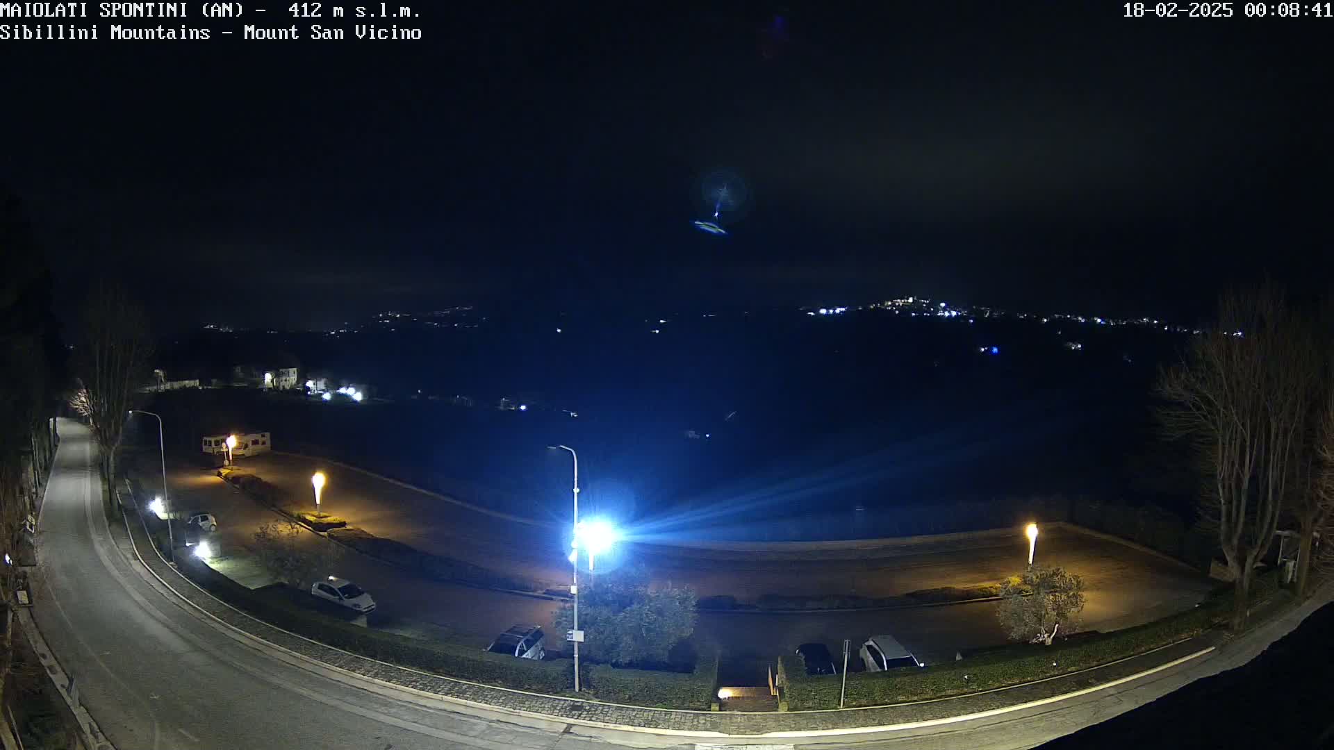 A nighttime, high-angle view of a paved road curving through a parking lot with several cars, illuminated by streetlights, against a backdrop of distant city lights and a dark sky.