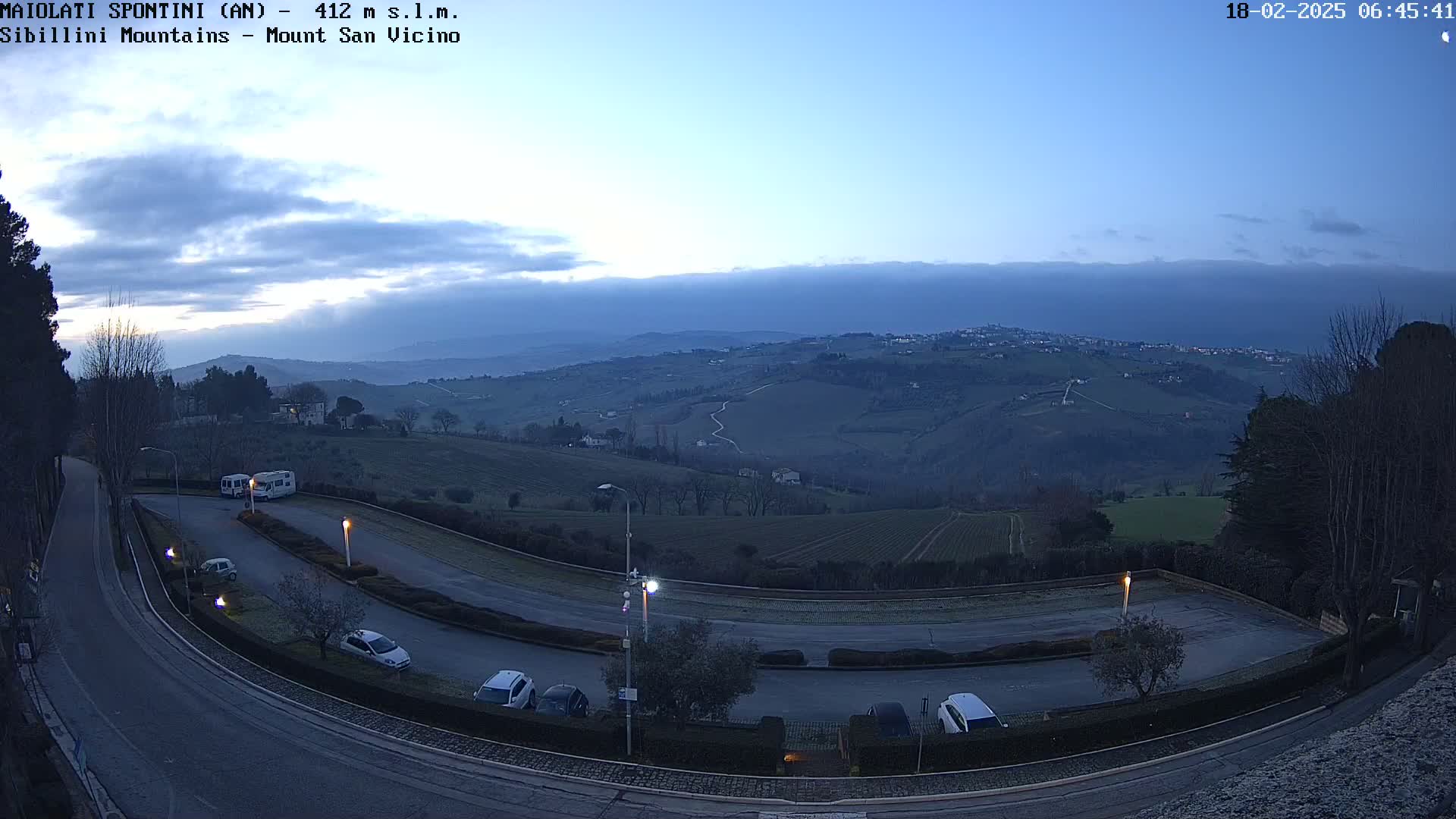A high-angle, twilight view shows a parking lot with several cars and a camper van, overlooking rolling hills and a distant town under a mostly clear, dusky sky.