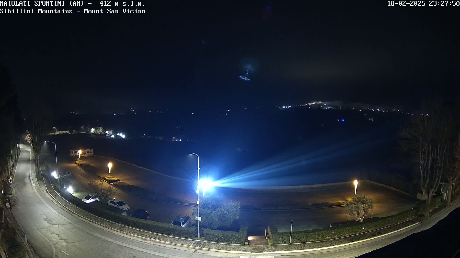 A nighttime, slightly hazy view of a curving road with parked cars and distant city lights, illuminated by bright streetlights.
