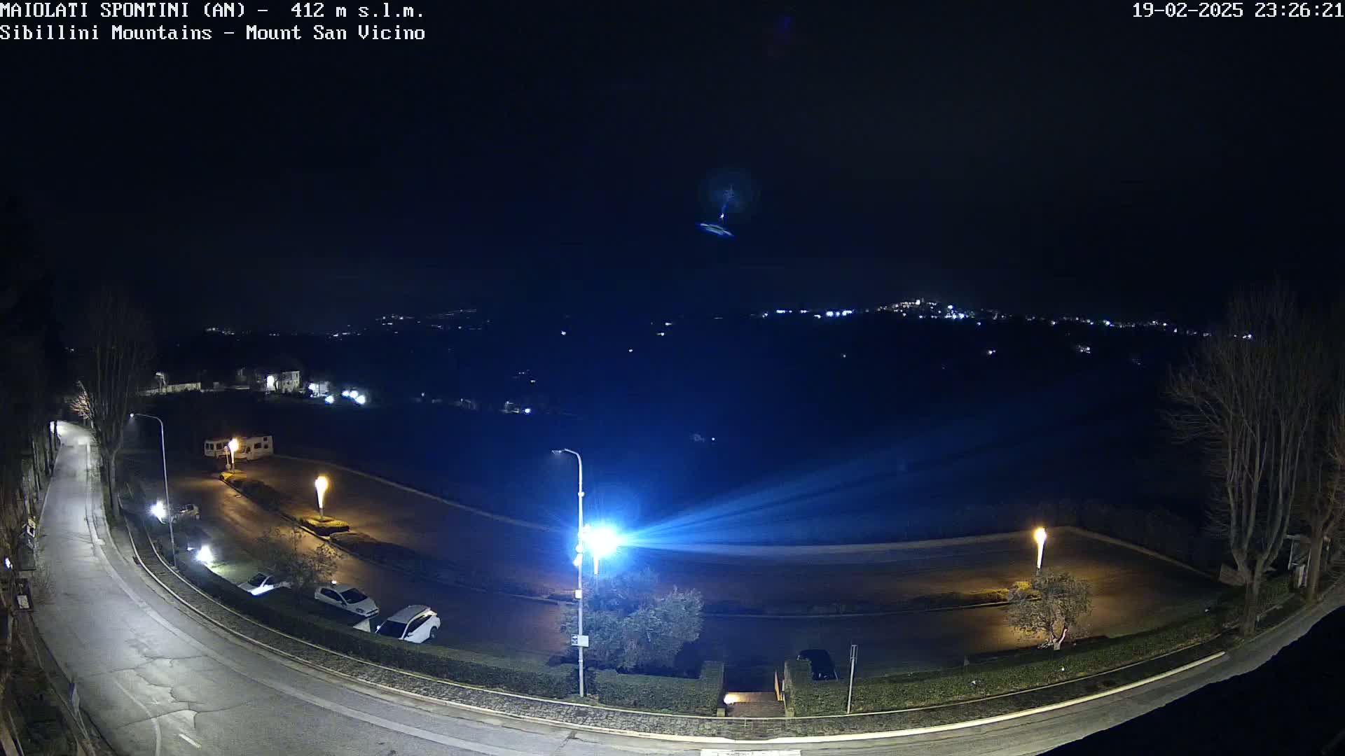 A nighttime view of a paved road curving through a parking lot, illuminated by streetlights, with a distant town visible under a clear, dark sky.
