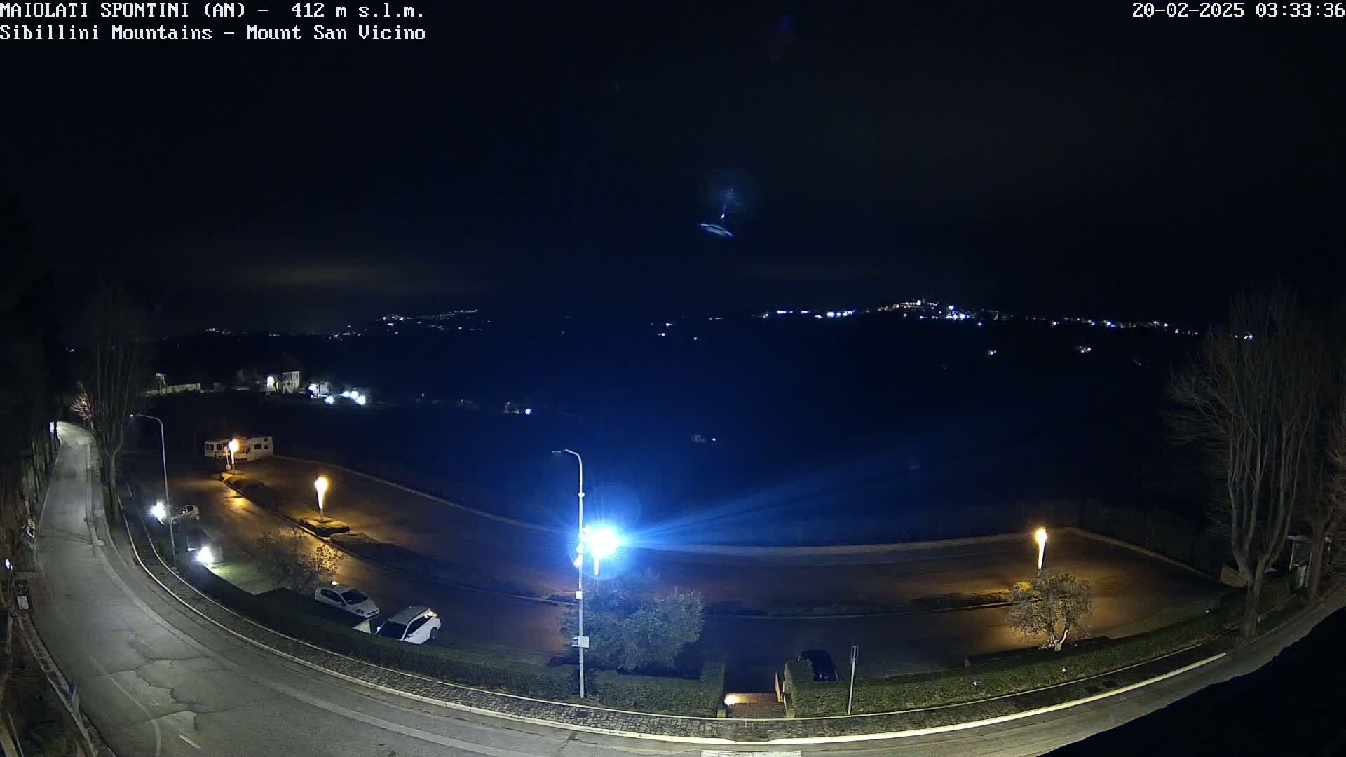 A nighttime, overhead view of a curving road with parked cars and streetlights, overlooking a distant, dimly lit town under a dark sky.