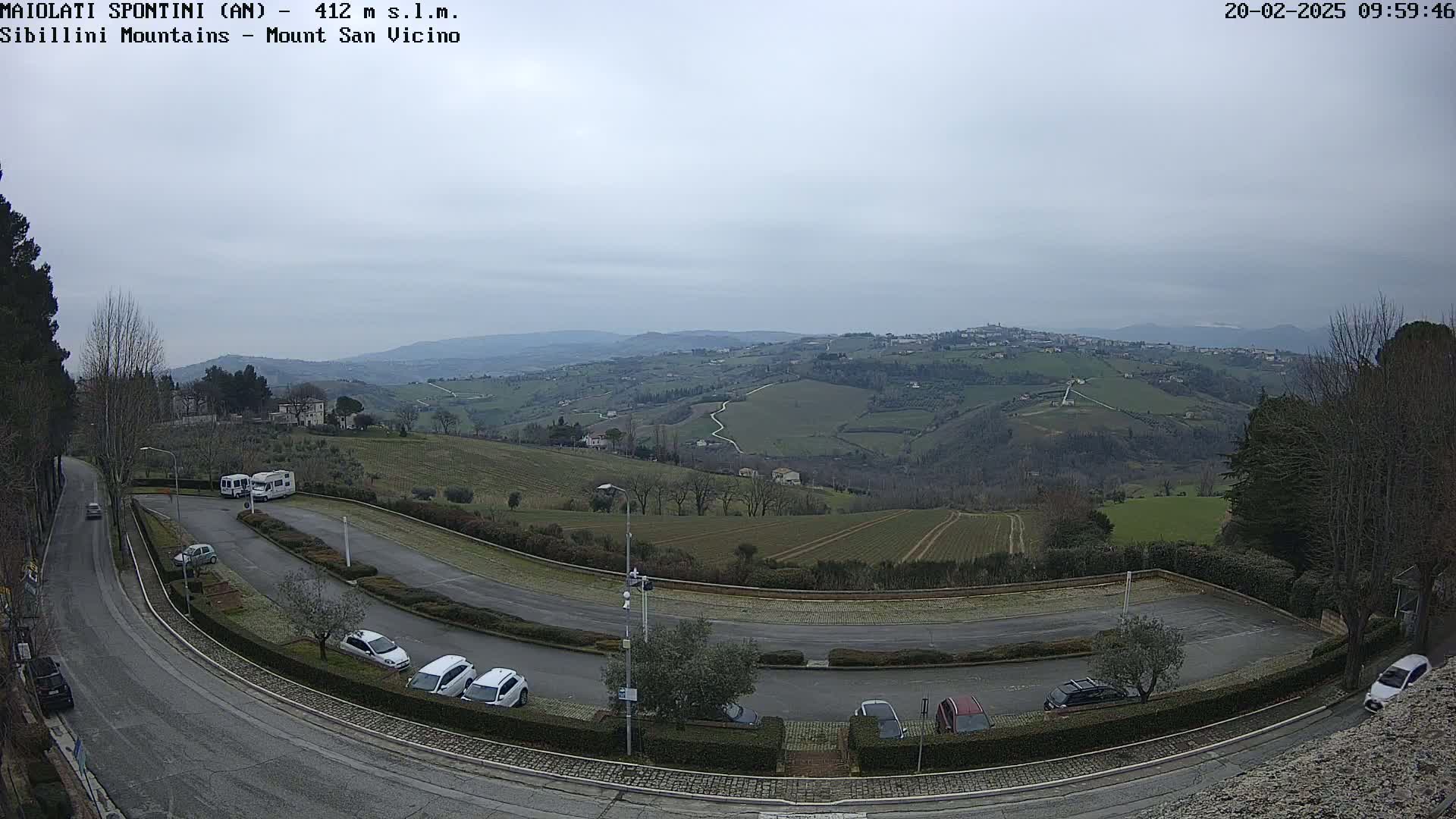A mostly cloudy day reveals a view of a parking lot, a curving road, and rolling hills dotted with buildings and farmland in the distance.