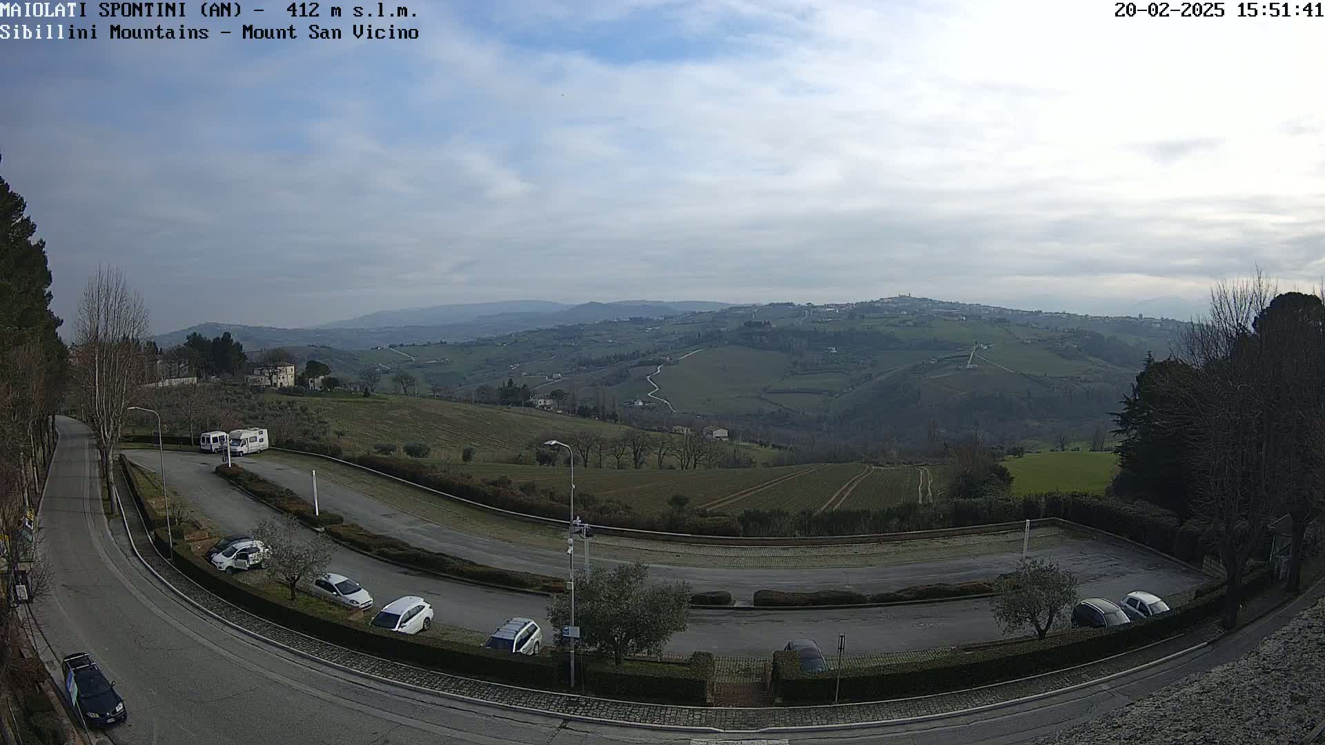 An aerial view shows a parking lot with several cars and two campervans, curving roads, and a hilly landscape under a mostly cloudy sky.