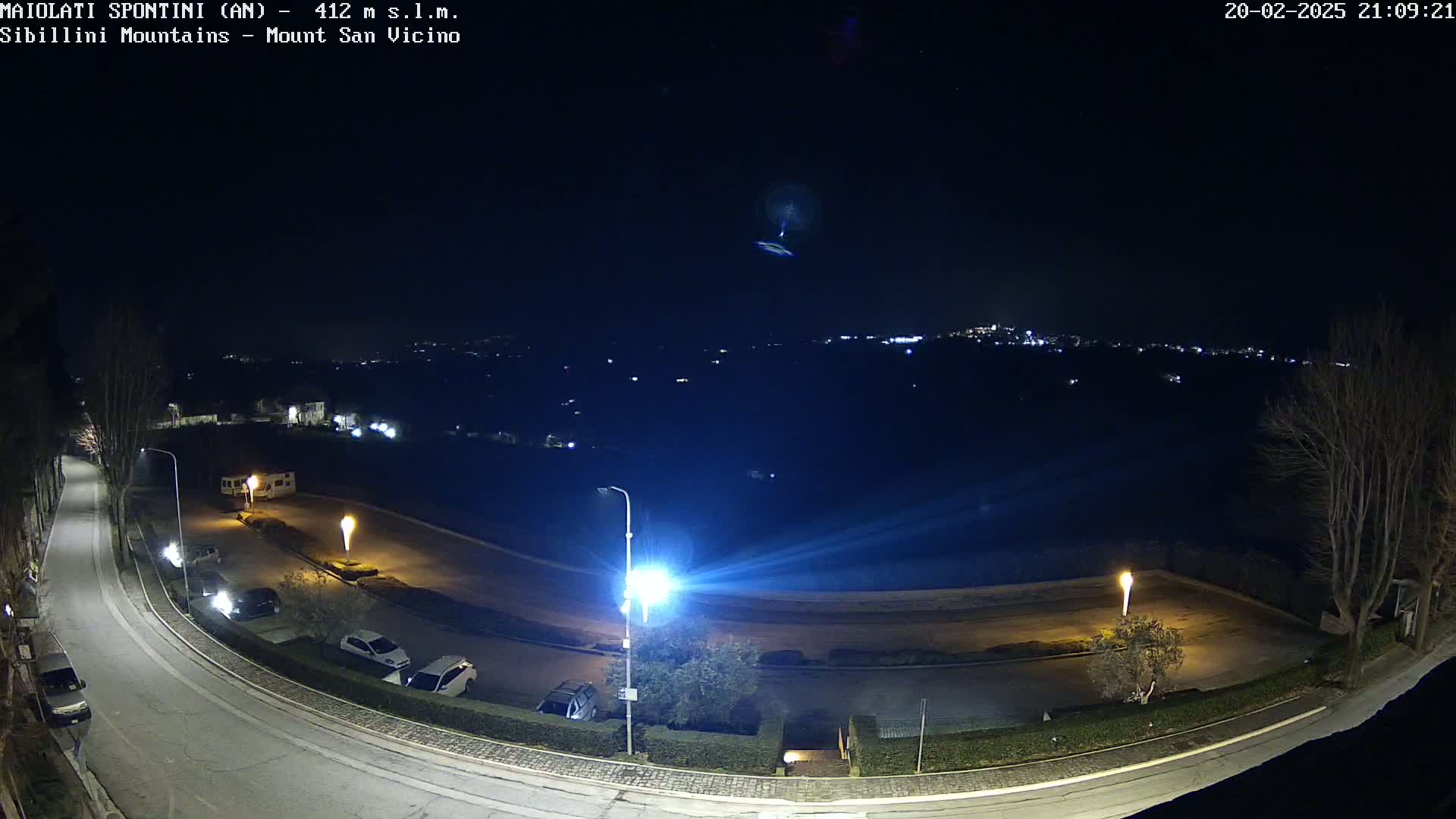 A nighttime aerial view of a curving road with parked cars and streetlights, overlooking a distant town illuminated under a clear, dark sky.