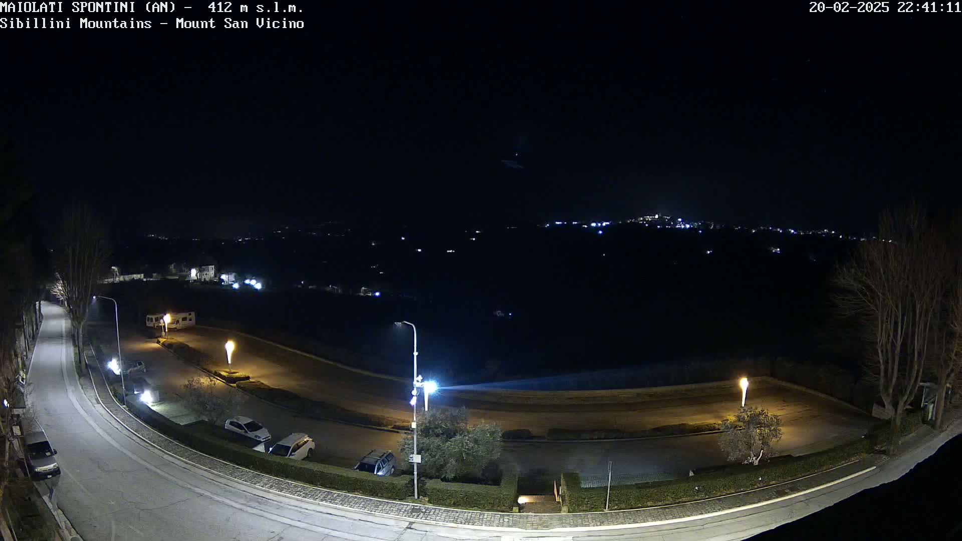 A nighttime, high-angle view shows a curving road with parked cars and some buildings, illuminated by streetlights, against a dark background with distant city lights visible on a hill.