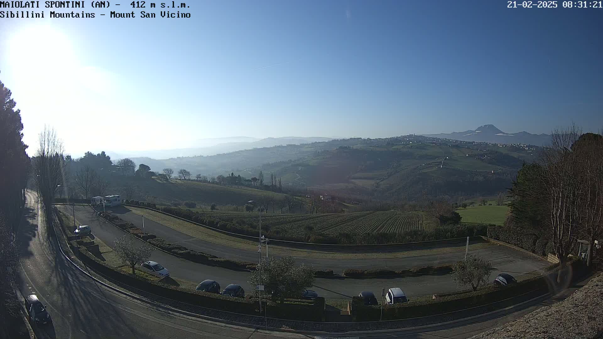 A sunny, hazy day reveals a winding road curving through a parking lot, with cars parked and a distant view of rolling hills and mountains under a clear sky.