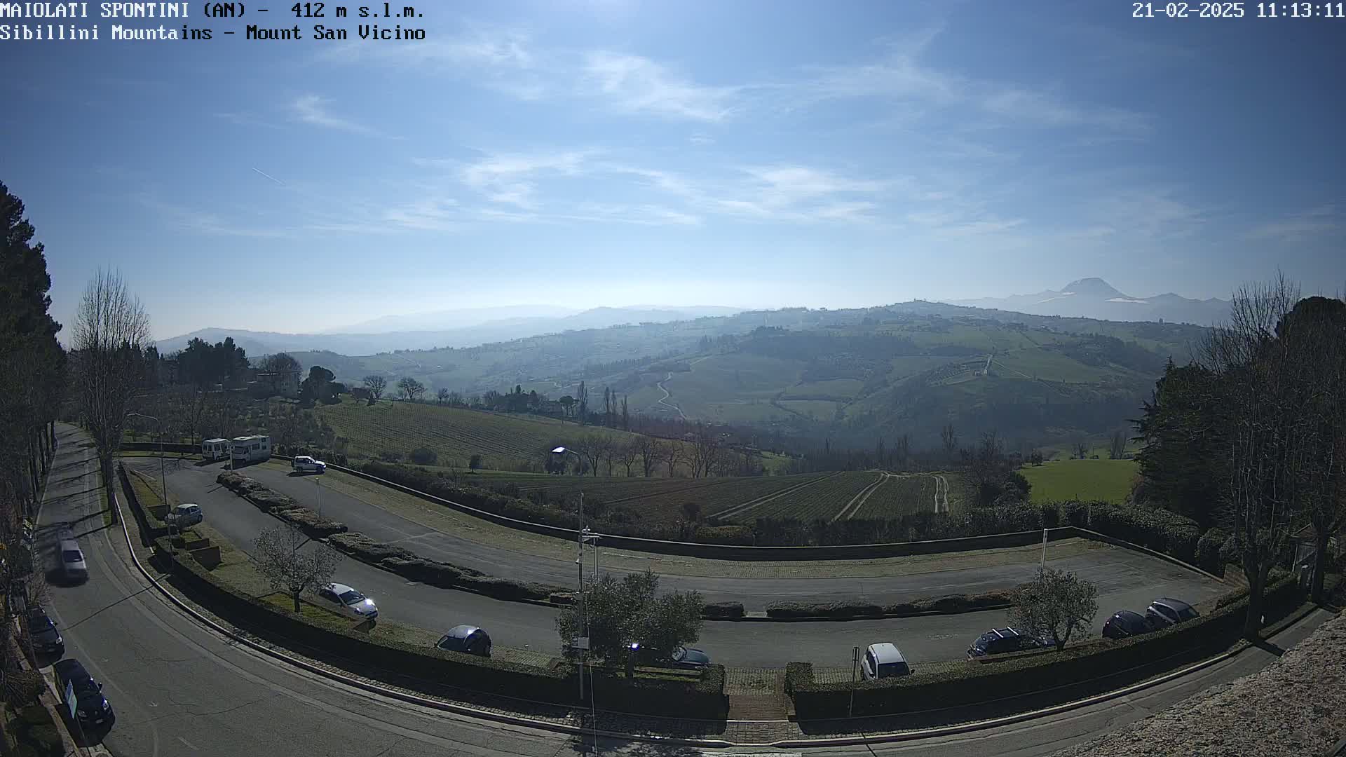 A sunny, clear day reveals a hilly landscape with vineyards, a parking lot filled with cars, and distant mountains under a bright blue sky.