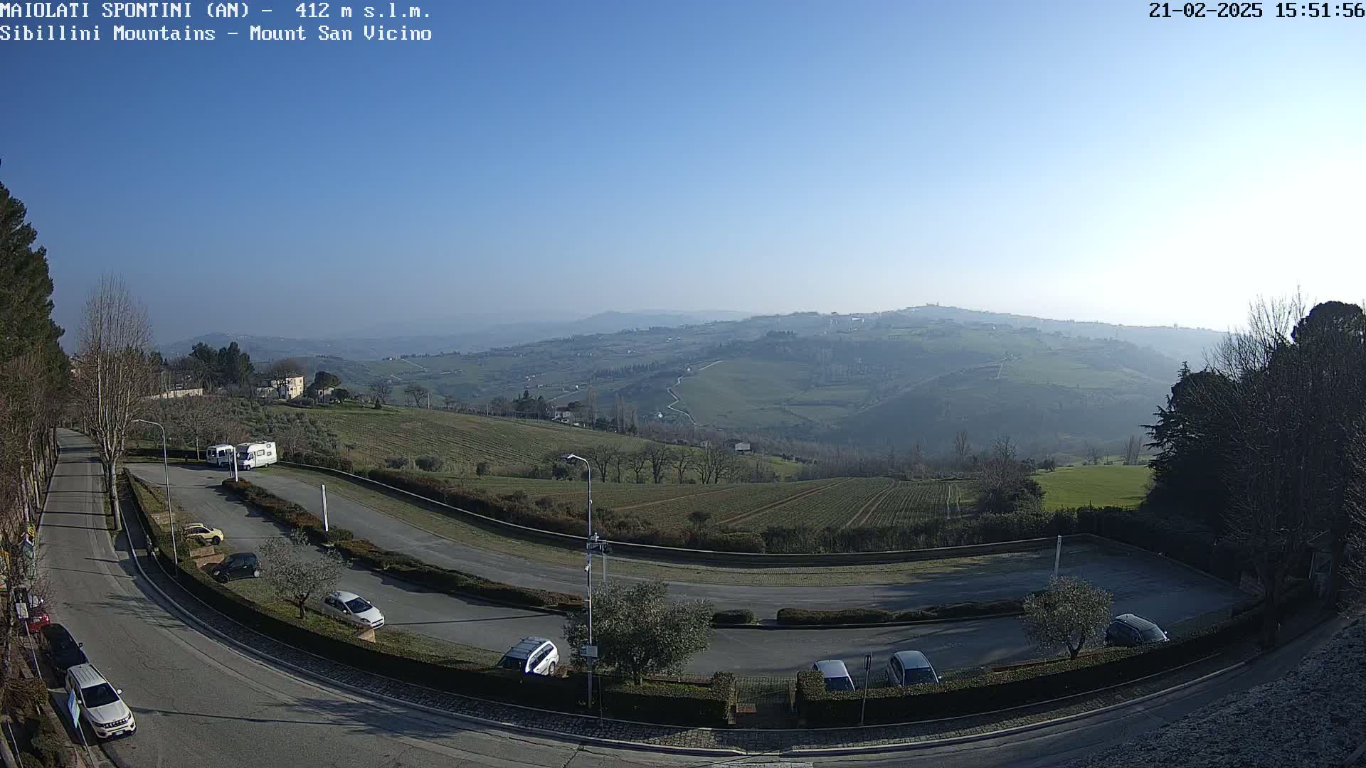 A mostly clear, sunny day overlooks a hilly landscape with a parking lot and several cars near a road.