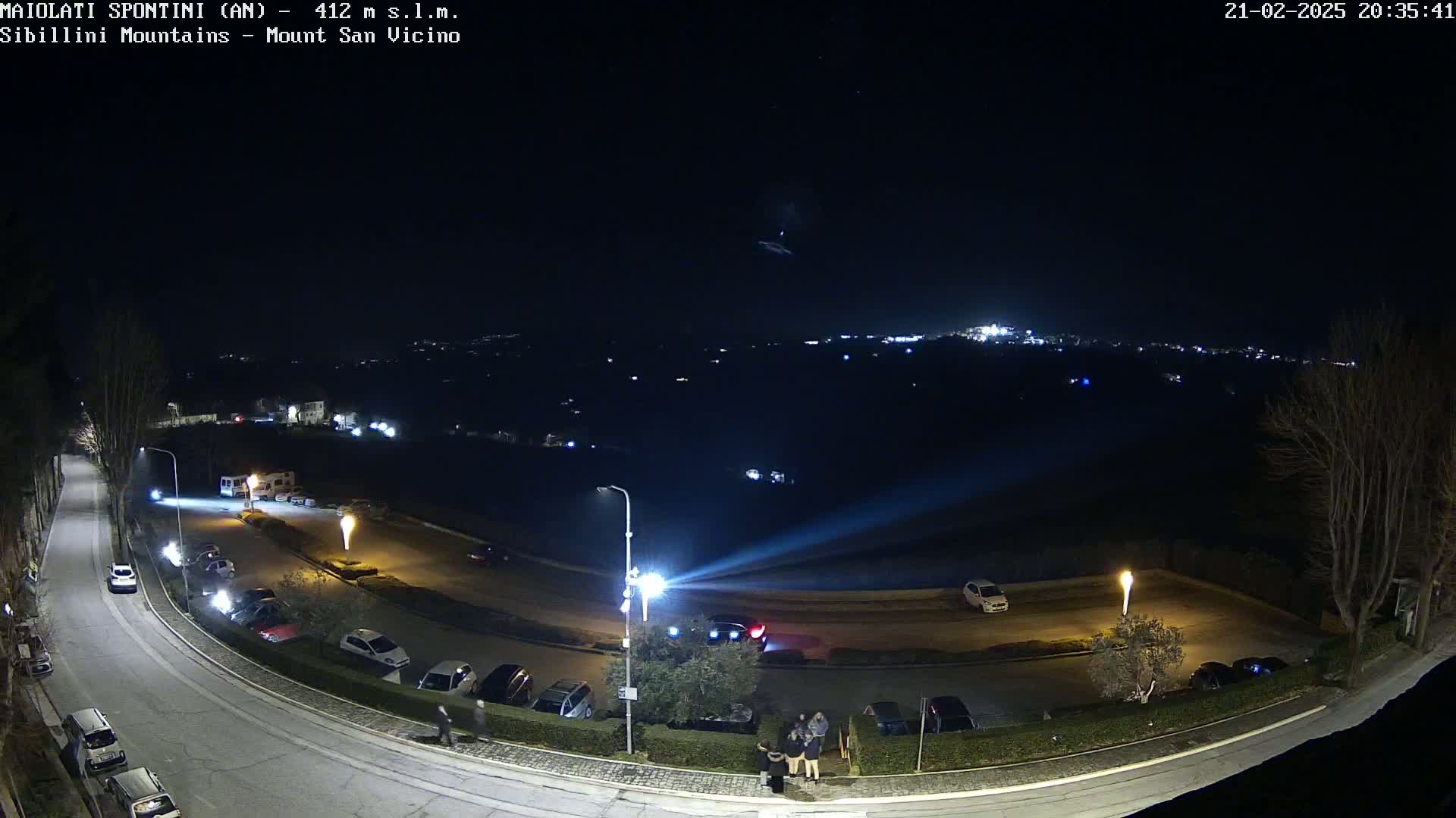 A nighttime aerial view of a parking lot and roadway with several cars parked, a few people standing near a building, and distant city lights under a clear, dark sky.