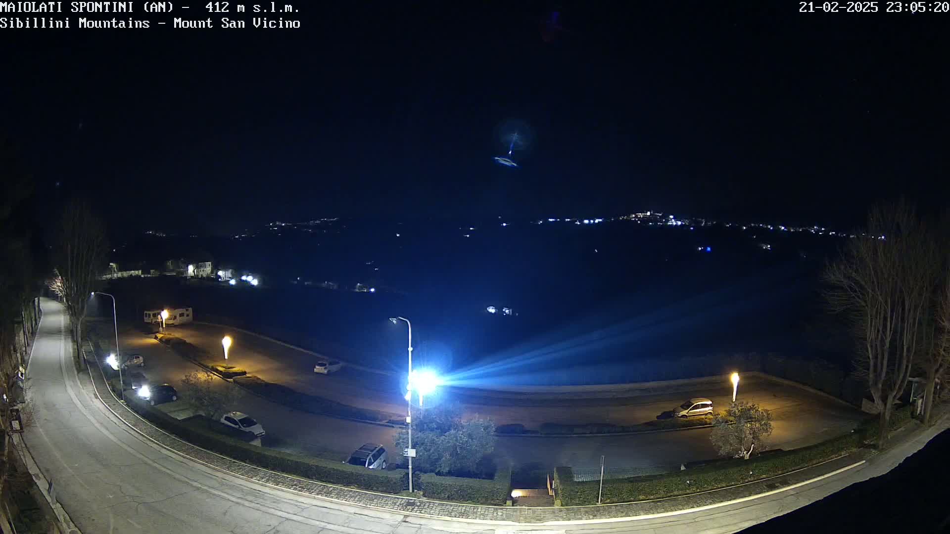 A nighttime aerial view of a curving road with parked cars and illuminated streetlights, overlooking a distant town under a clear, dark sky.