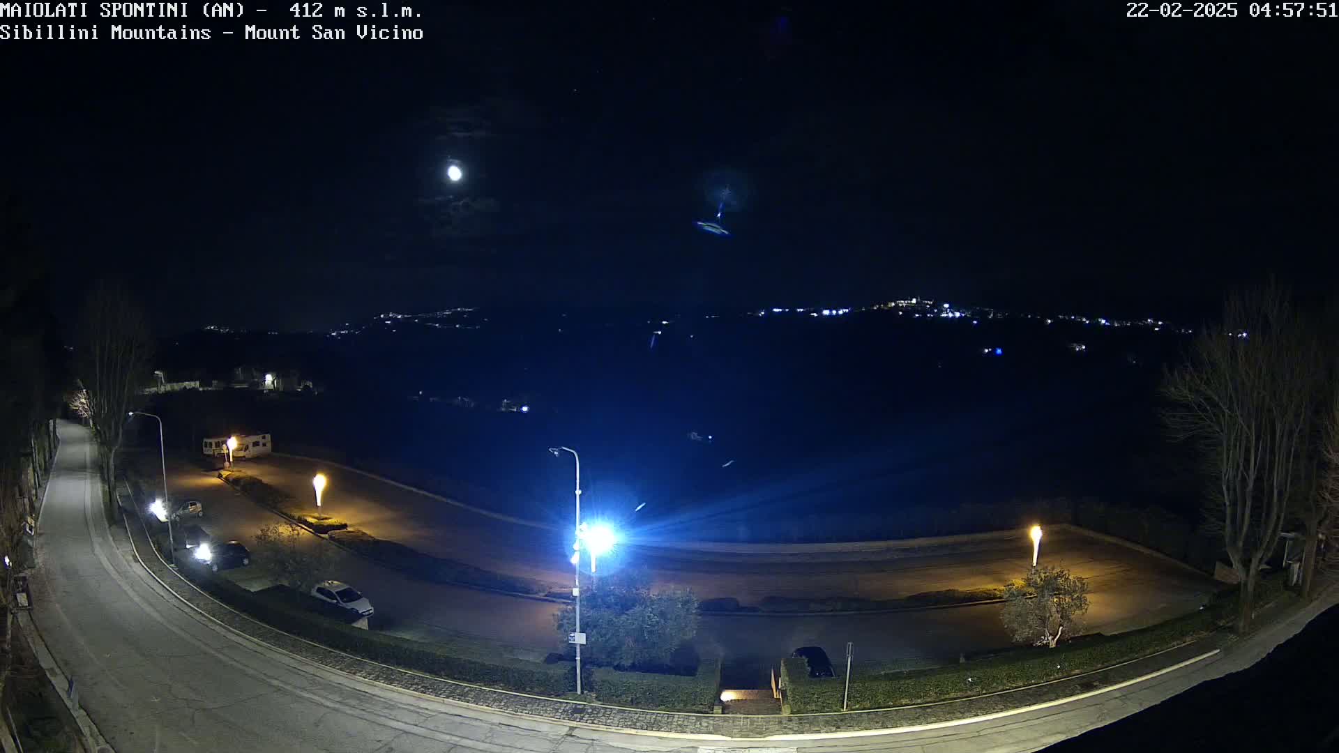 A nighttime, high-angle view of a curved road with parked cars, illuminated by streetlights, overlooking a distant town nestled in a hilly landscape under a moonlit sky.