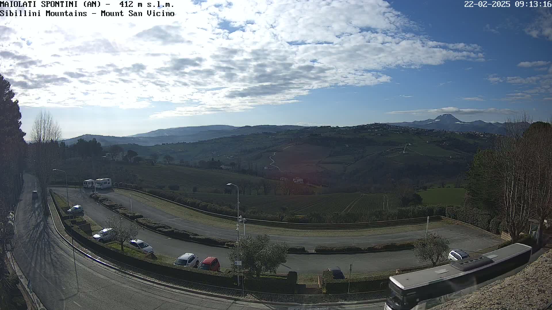 A partly cloudy blue sky overlooks a hilly landscape with vineyards and a parking lot containing several cars and buses.