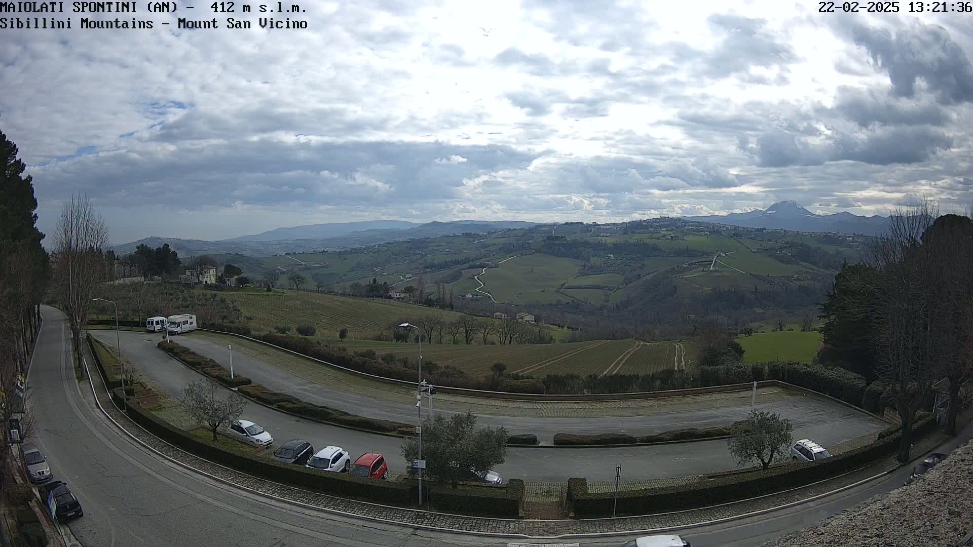 A paved road curves through a parking lot overlooking a hilly landscape under a partly cloudy sky, with several cars parked and distant mountains visible in the background.