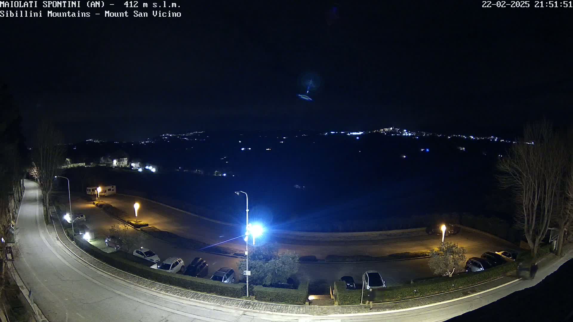 A nighttime, high-angle view of a curved road with parked cars and a distant town illuminated under a clear, dark sky.