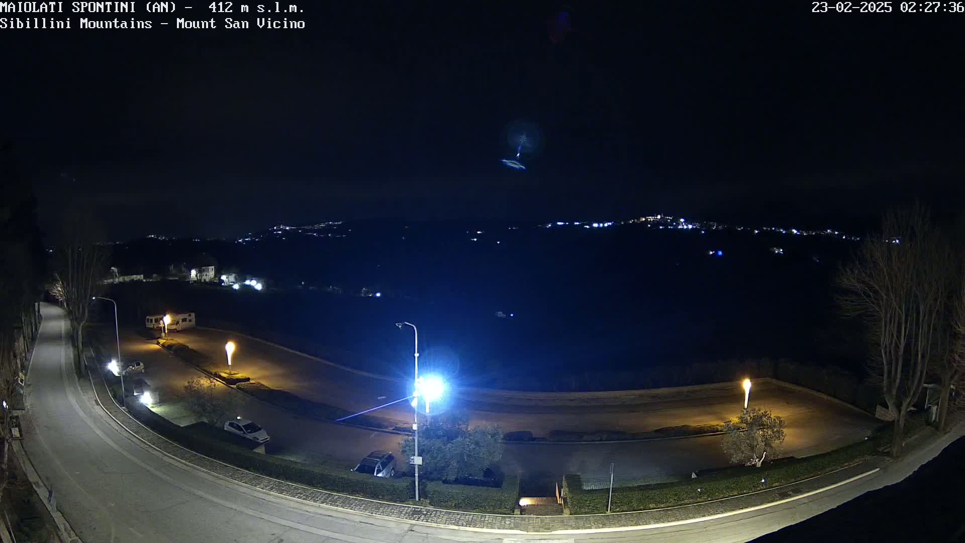 A nighttime, high-angle view of a curving road illuminated by streetlights, with a few parked cars, and a distant, dimly lit town nestled in the hills under a clear, dark sky.