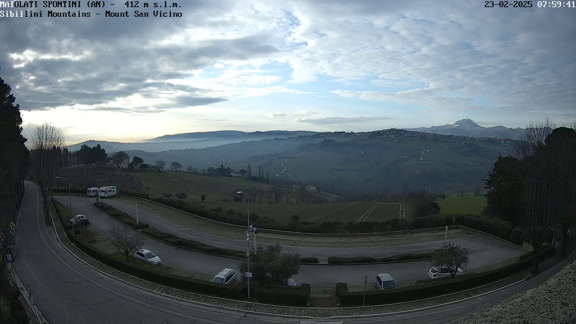A winding road with parked cars overlooks a valley shrouded in morning mist, under a partly cloudy sky, with distant mountains visible in the background.