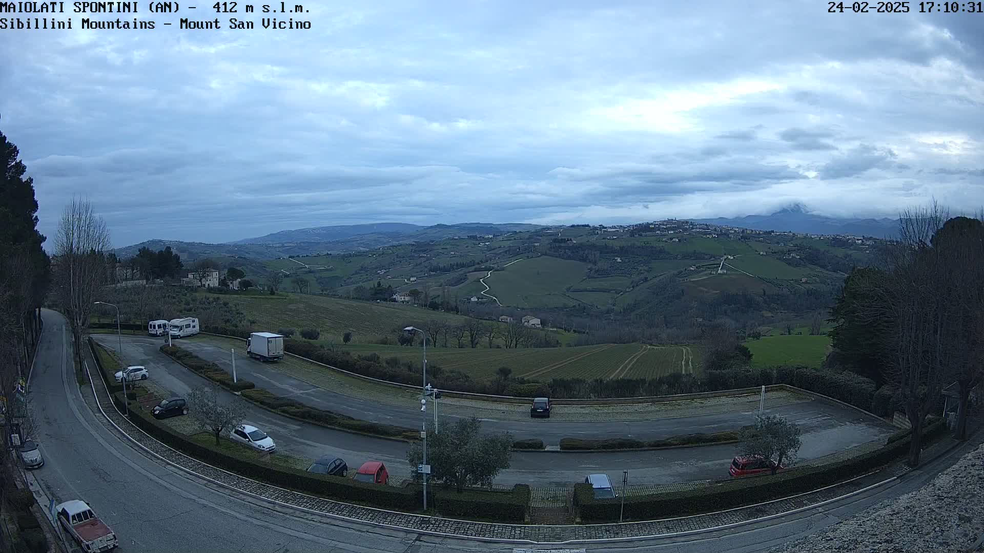 A parking lot with several cars and trucks overlooks a sprawling, hilly landscape under a cloudy sky.