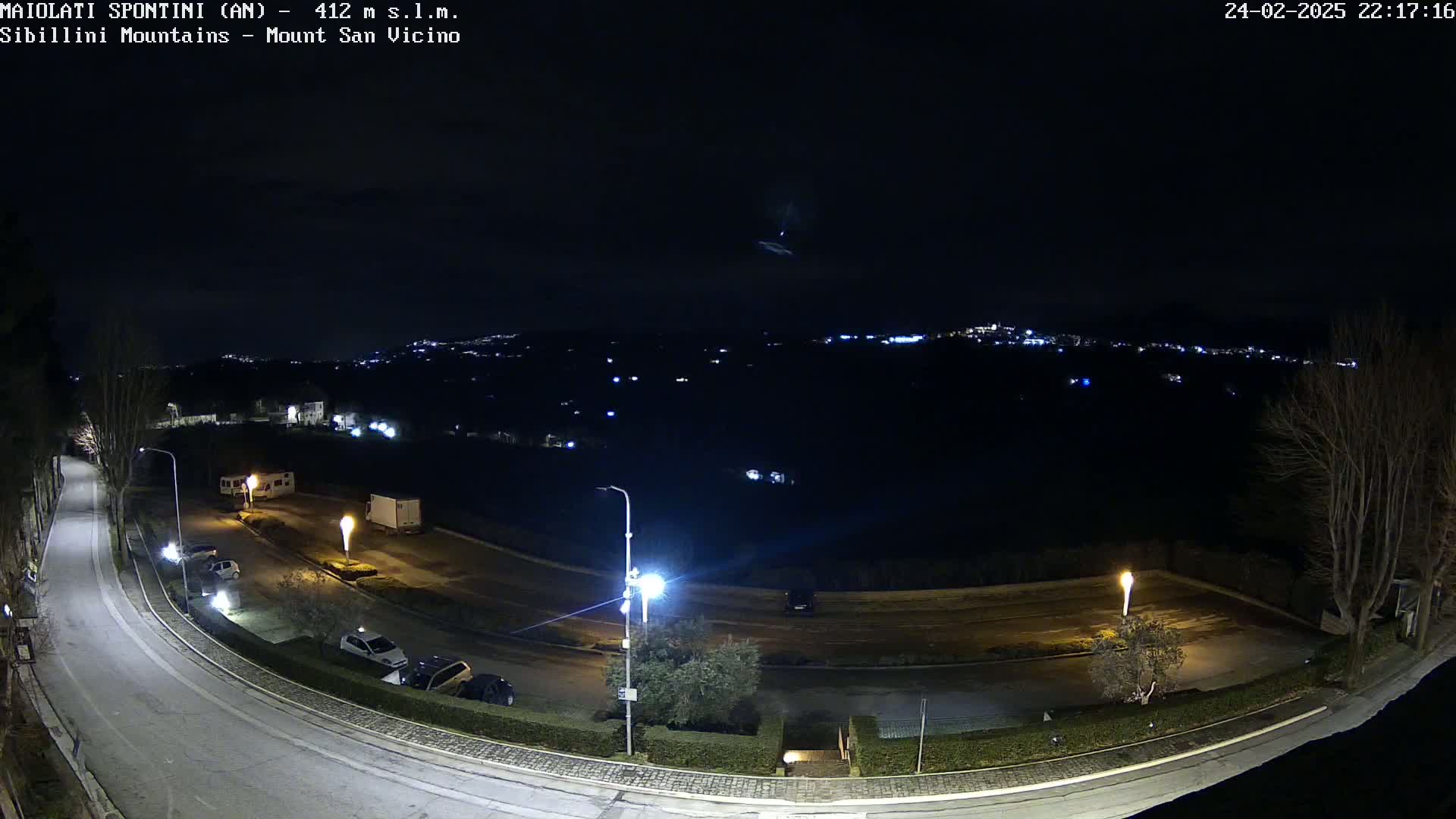 A nighttime, high-angle view of a curved road with parked cars and a few vehicles, leading to a parking area next to a dark field under a clear night sky with distant city lights.