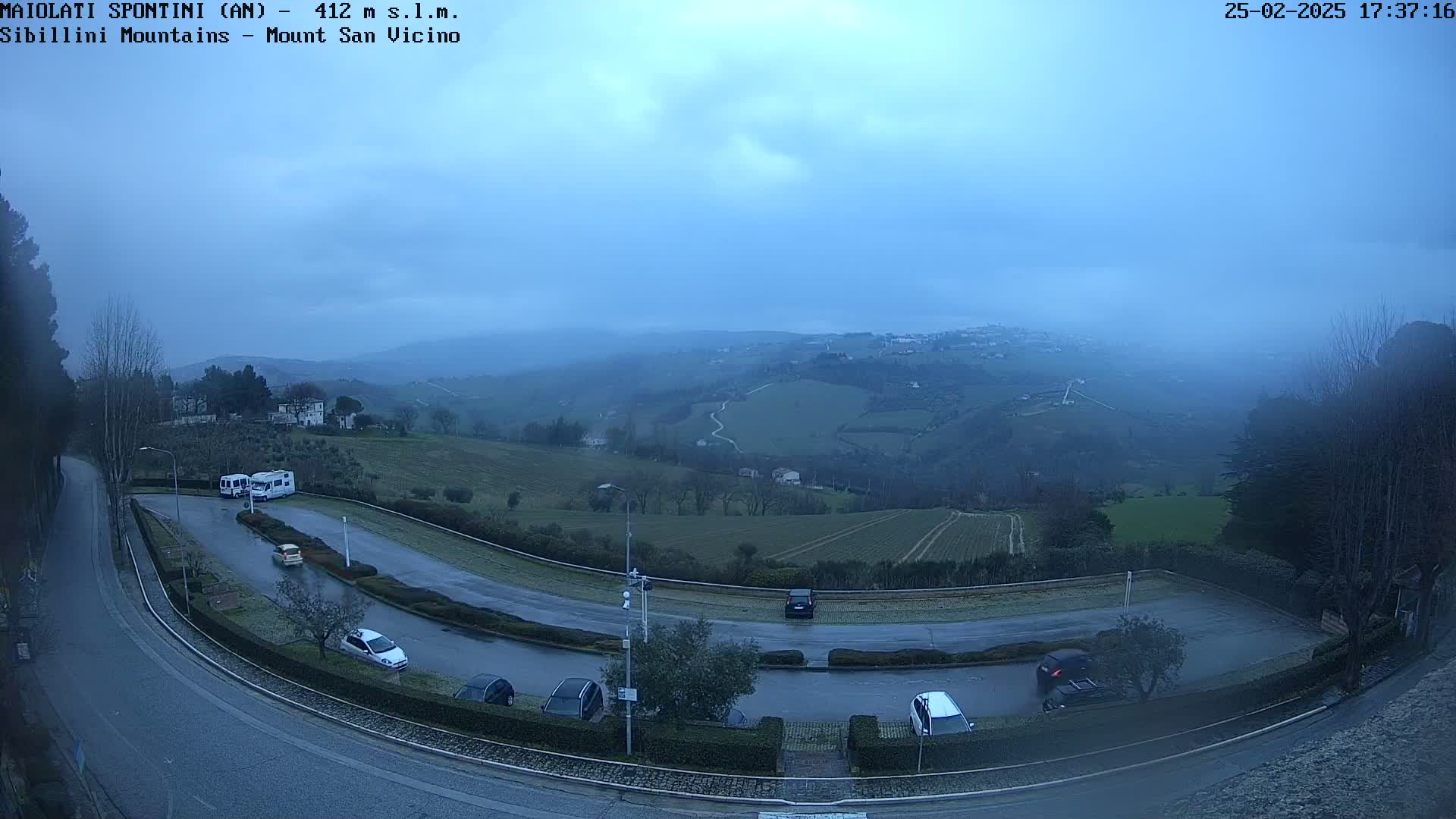 A high-angle, long shot shows a parking lot with several cars and two RVs next to a winding road, overlooking a misty, hilly landscape under a dusky blue sky.