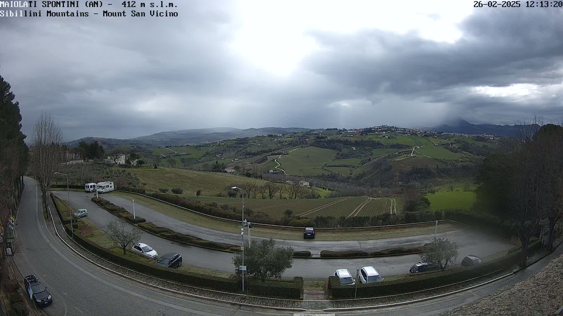 A parking lot with several cars and RVs overlooks a hilly, green landscape under a cloudy, overcast sky with light rain.