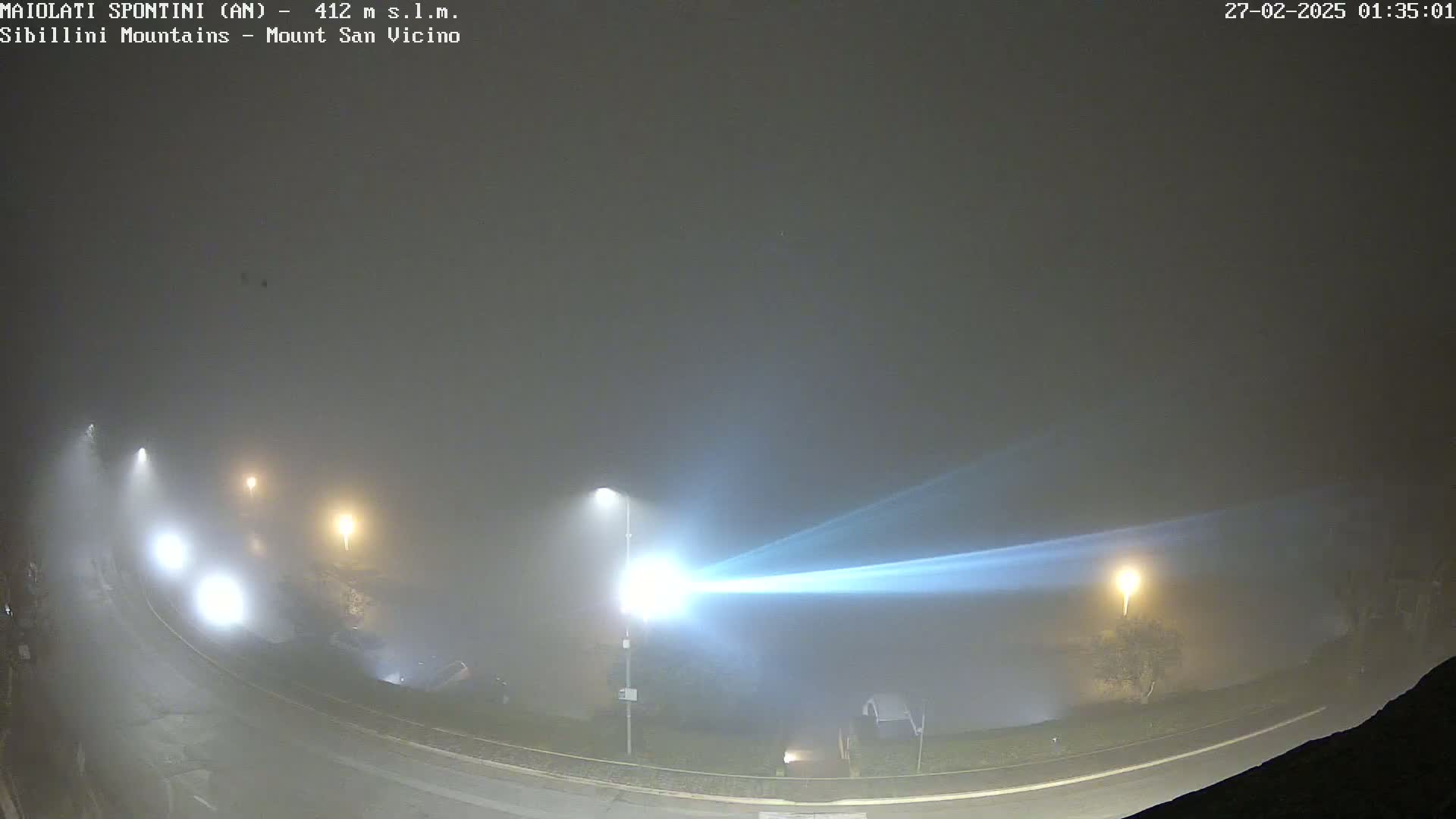 A foggy night scene shows a curved road with several cars and streetlights, their beams cutting through the thick fog.